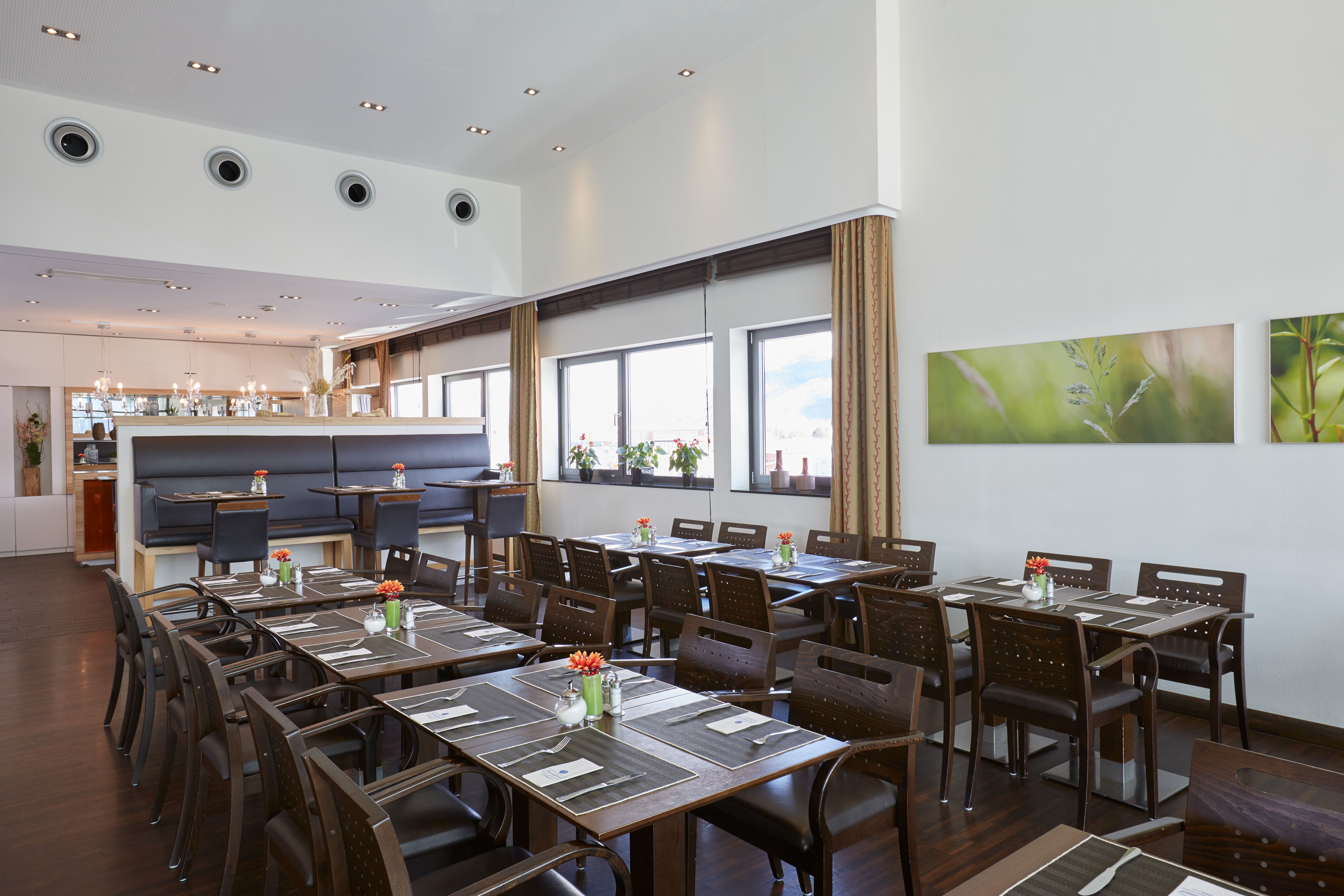 Helles, modernes Restaurant mit großen Fenstern, dunkelbraunen Holztischen und -stühlen, gedeckten Tischen und frischen Blumen. Freundliche Atmosphäre für Frühstück, Mittag- oder Abendessen in stilvollem Ambiente.