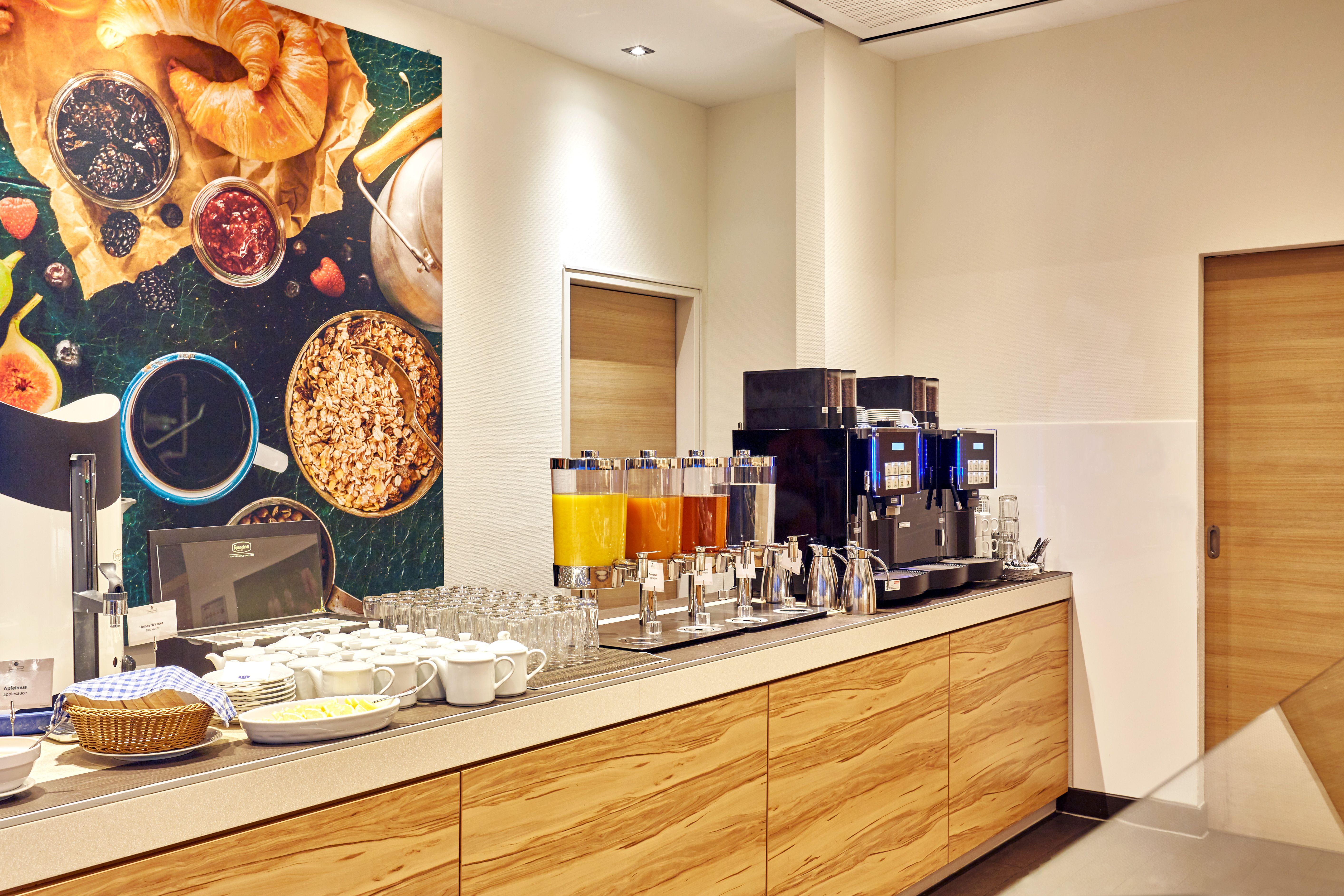 Frühstücksbereich mit Kaffeeautomat, Saftspendern, Gläsern, Tassen und Schalen für ein vielfältiges Buffet, freundliche Atmosphäre für einen gelungenen Start in den Tag.