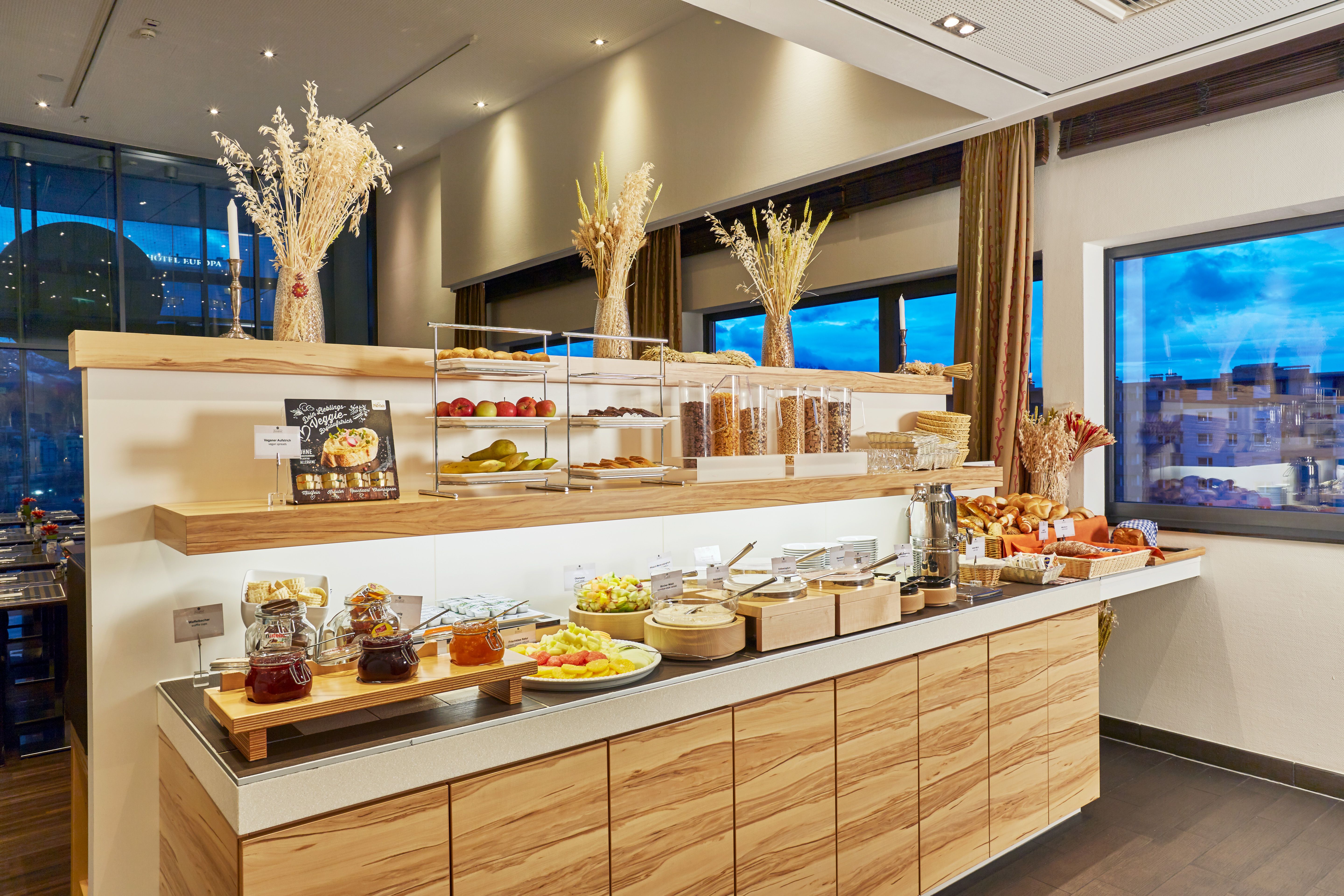 Buffet mit frisch angerichteten Frühstücksangeboten wie Obst, Müsli, Brot und Aufstrichen in einem hellen, modernen Raum mit Fenstern und natürlicher Dekoration.