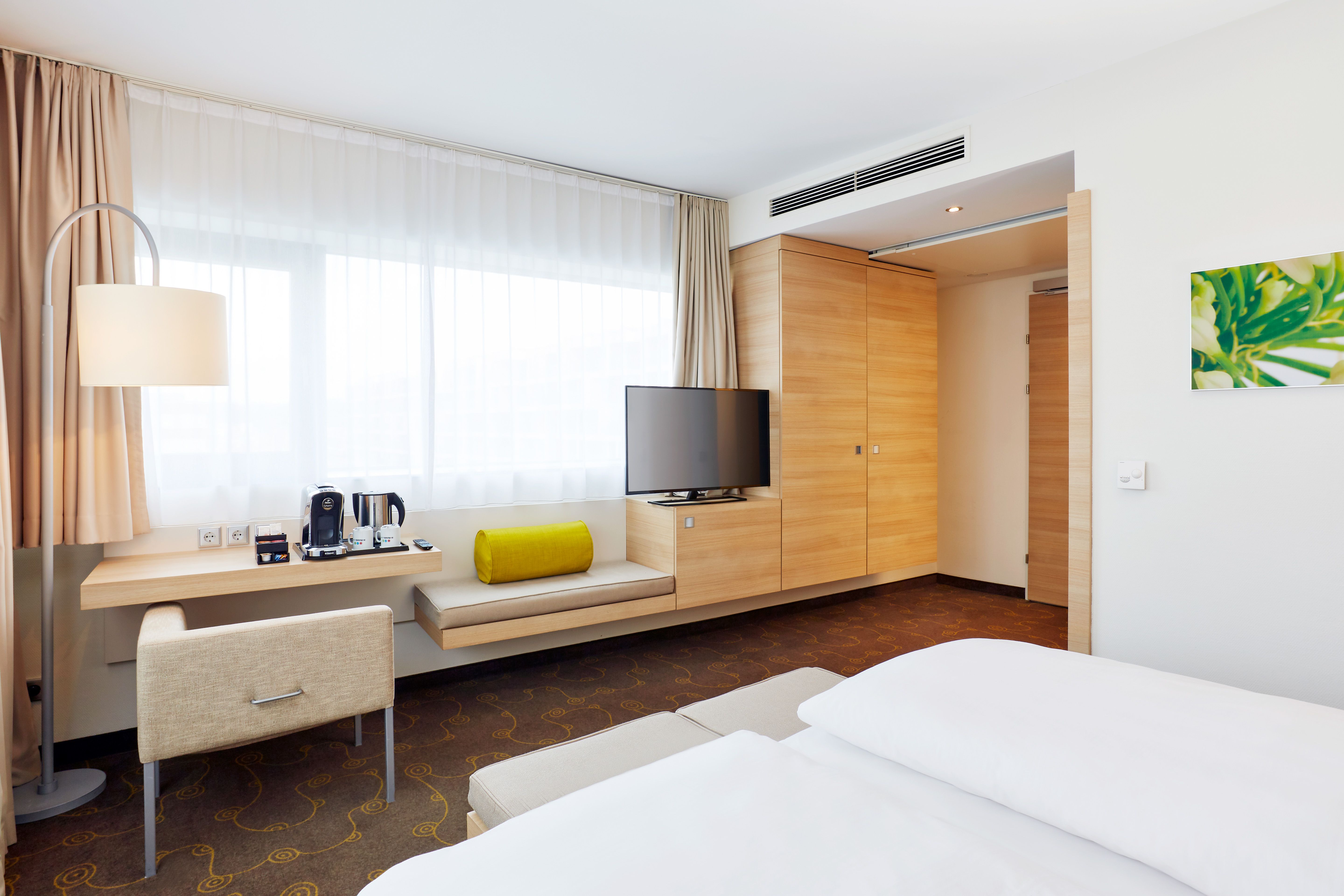 Modernes Hotelzimmer mit großem Fenster, hellem Tageslicht, gemütlichem Sitzbereich, Schreibtisch, Flachbildfernseher und Doppelbett. Natürliche Holzmöbel und dezente Dekoration schaffen eine entspannte Atmosphäre.