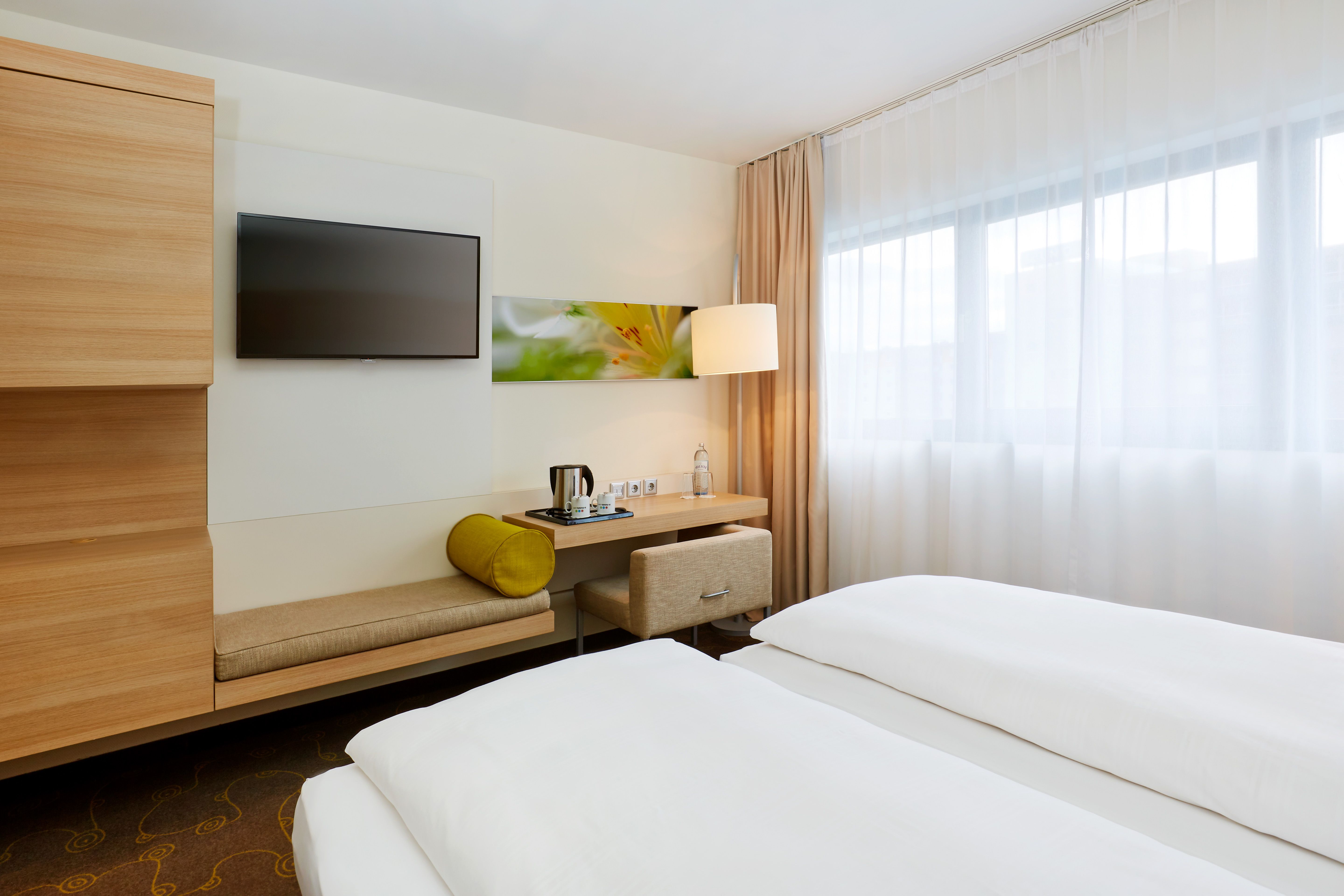 Modern eingerichtetes Hotelzimmer mit großem Fenster, zwei Betten, Sitzbank, Schreibtisch mit Wasserflasche und Teekanne sowie Fernseher an der Wand. Helle, freundliche Atmosphäre.