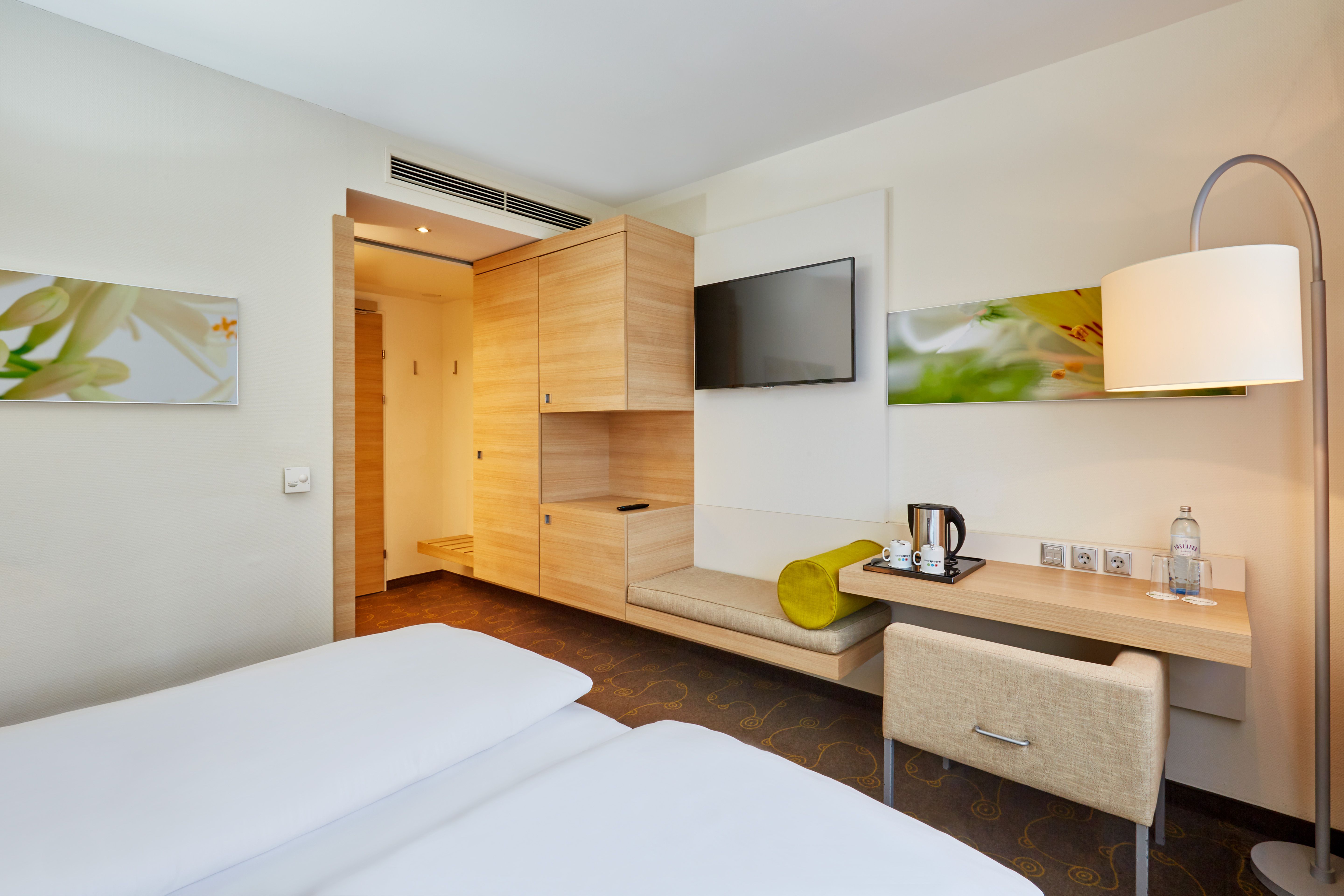 Helles, modernes Hotelzimmer mit Doppelbett, Holzmöbeln, Flachbildfernseher, Schreibtisch mit Wasserkocher und Flasche Wasser, sowie stimmungsvoller Beleuchtung. Angenehme Atmosphäre für komfortablen Aufenthalt.