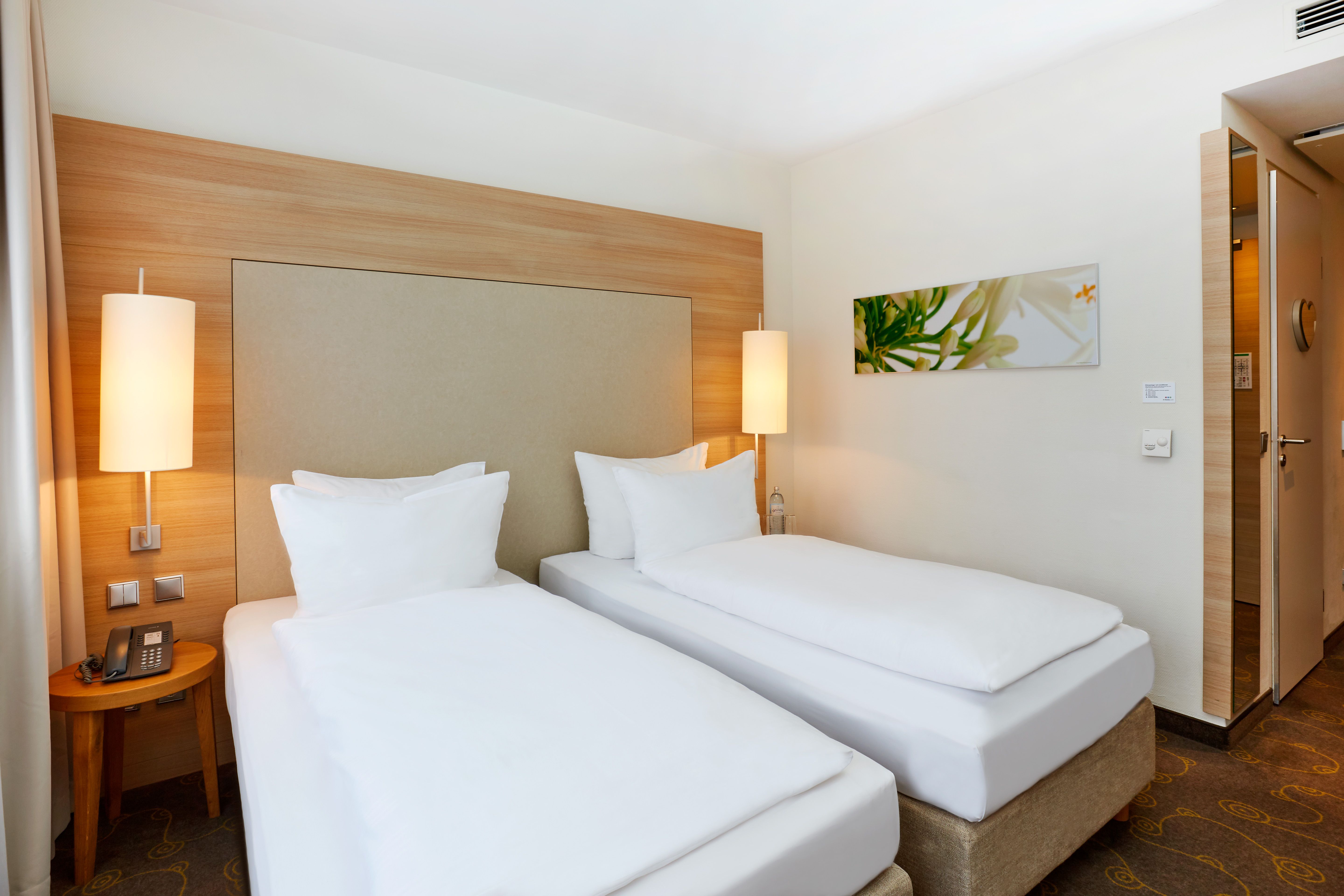 Helles Hotelzimmer mit zwei Einzelbetten, weißen Bettwäsche, warmem Holzdesign, zwei Nachttischlampen und modernem Wandbild für eine gemütliche und entspannte Atmosphäre.