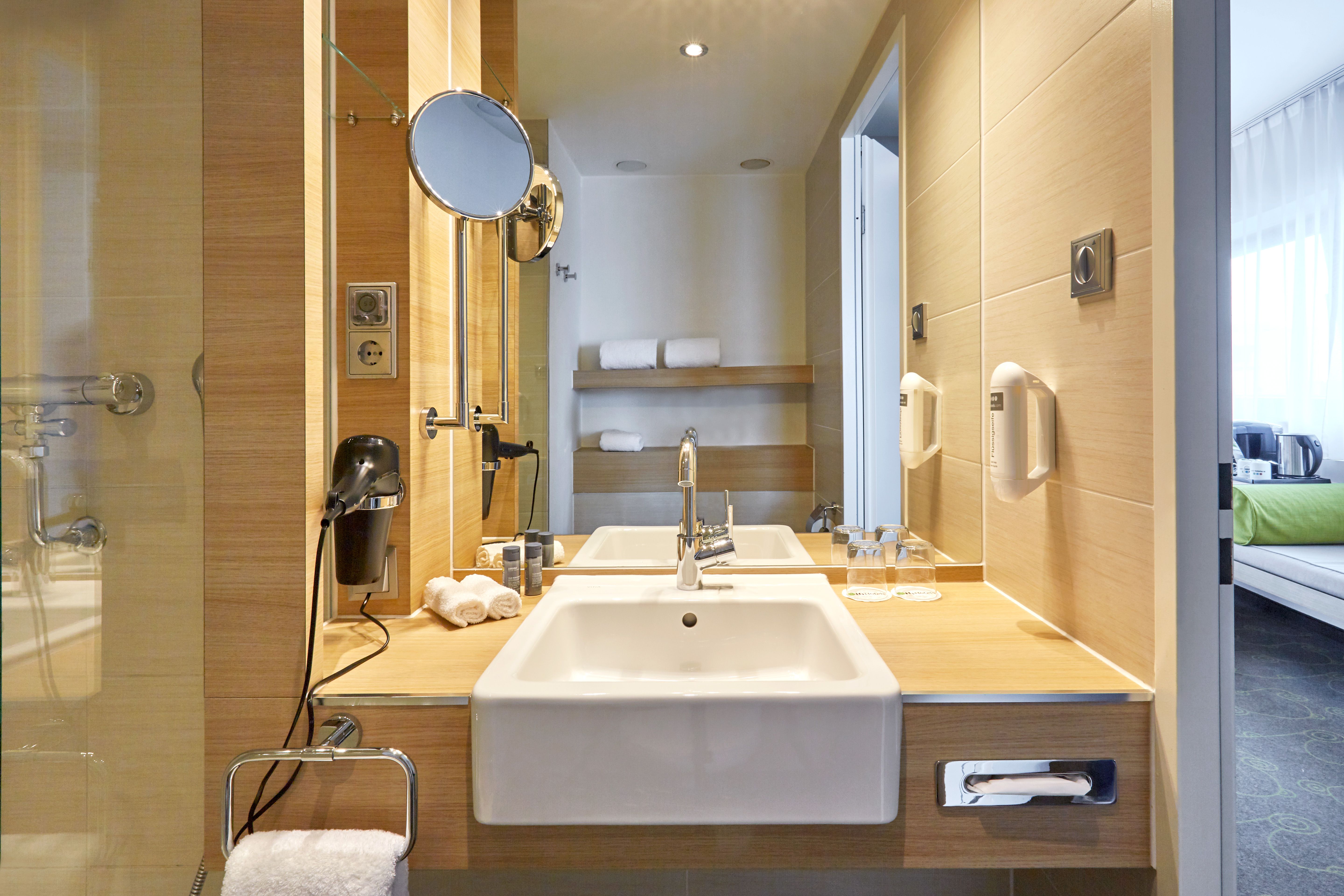 Modernes Badezimmer mit weißem Waschbecken, großem Spiegel, Holzarbeitsplatte und klarer Beleuchtung. Im Hintergrund sind Handtücher ordentlich auf Regalen verstaut und ein angrenzendes Gästezimmer teilweise sichtbar.