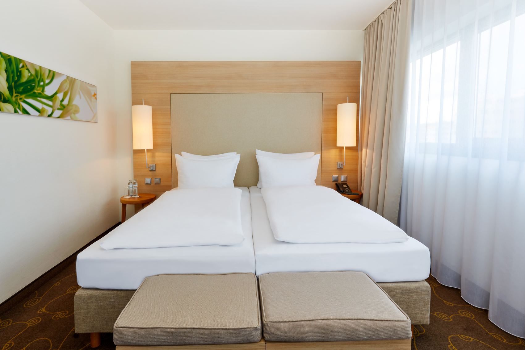 Modernes Hotelzimmer mit zwei einzelnen Betten, hellen Holzakzenten, großen Fenstern mit Vorhängen und sanfter Beleuchtung für eine ruhige und entspannte Atmosphäre.