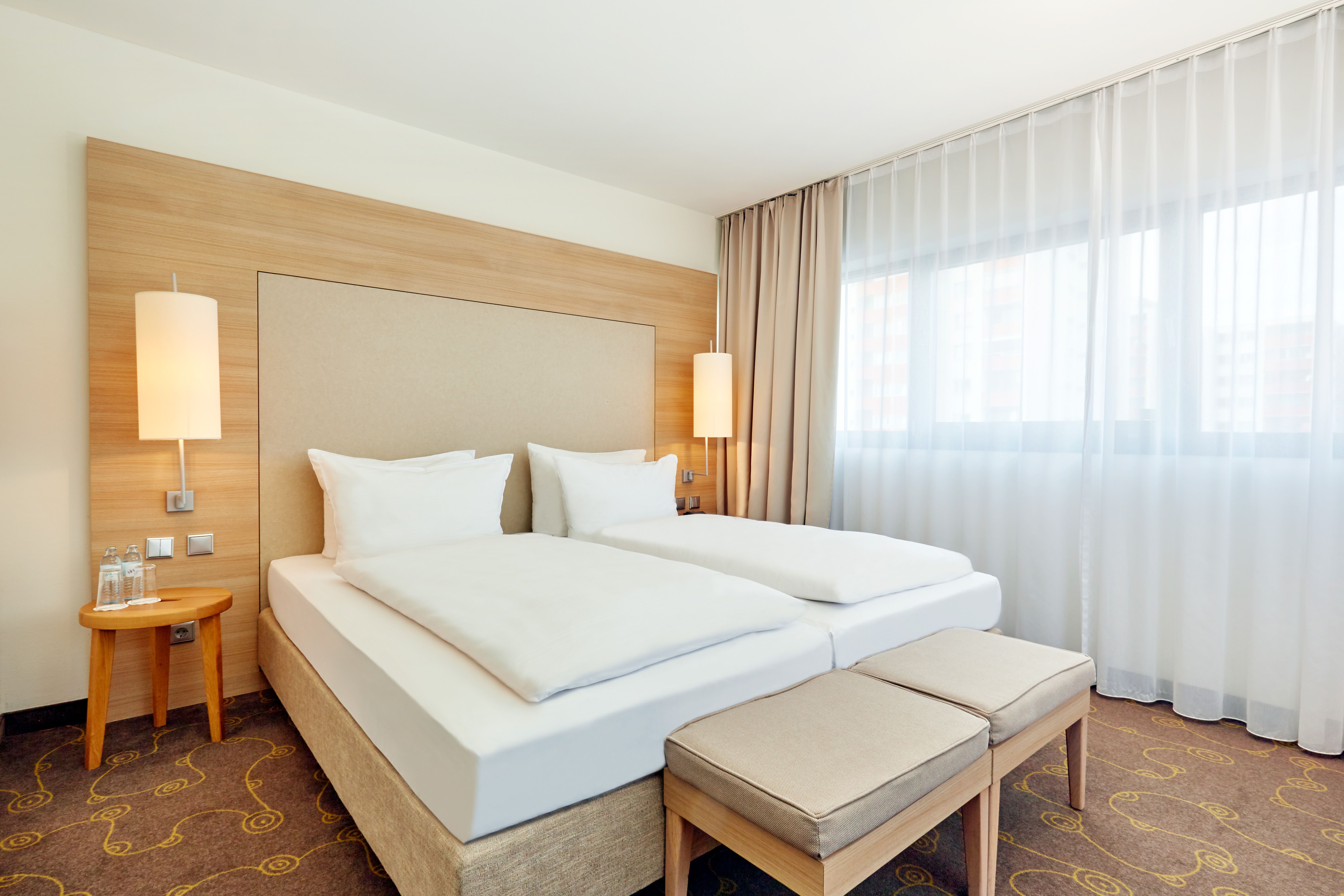 Modernes, helles Hotelzimmer mit zwei Einzelbetten, großen Fenstern mit transparenten Vorhängen, warmen Holzelementen und weicher Beleuchtung, das eine ruhige und einladende Atmosphäre zum Entspannen bietet.