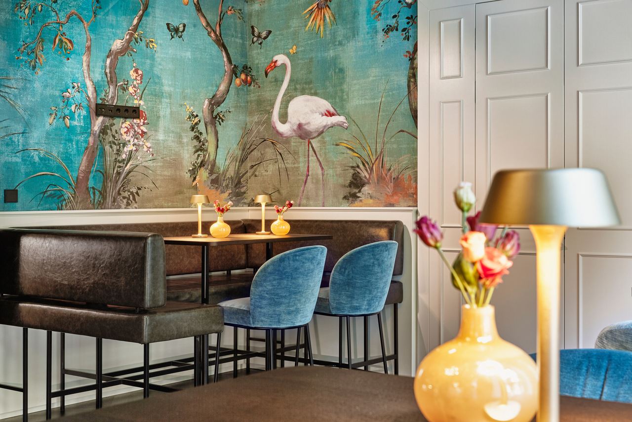 Gemütliche Sitzecke mit dunkelblauen Stühlen und brauner Sitzbank vor einer Wand mit farbenfrohem Flamingo-Tapetenmotiv, dekoriert mit Blumen und sanfter Beleuchtung für eine entspannte Atmosphäre.