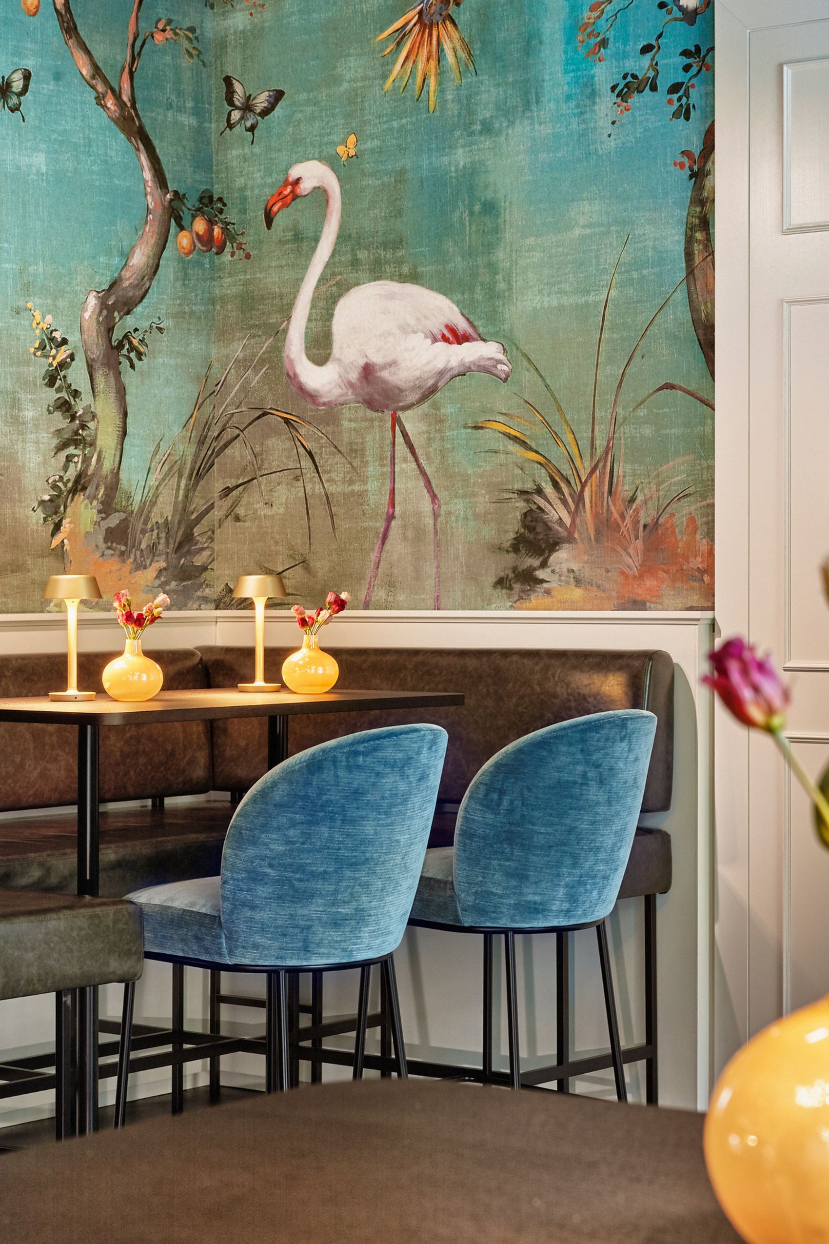 Gemütliche Sitzecke mit dunkelblauen Stühlen und brauner Sitzbank vor einer Wand mit farbenfrohem Flamingo-Tapetenmotiv, dekoriert mit Blumen und sanfter Beleuchtung für eine entspannte Atmosphäre.