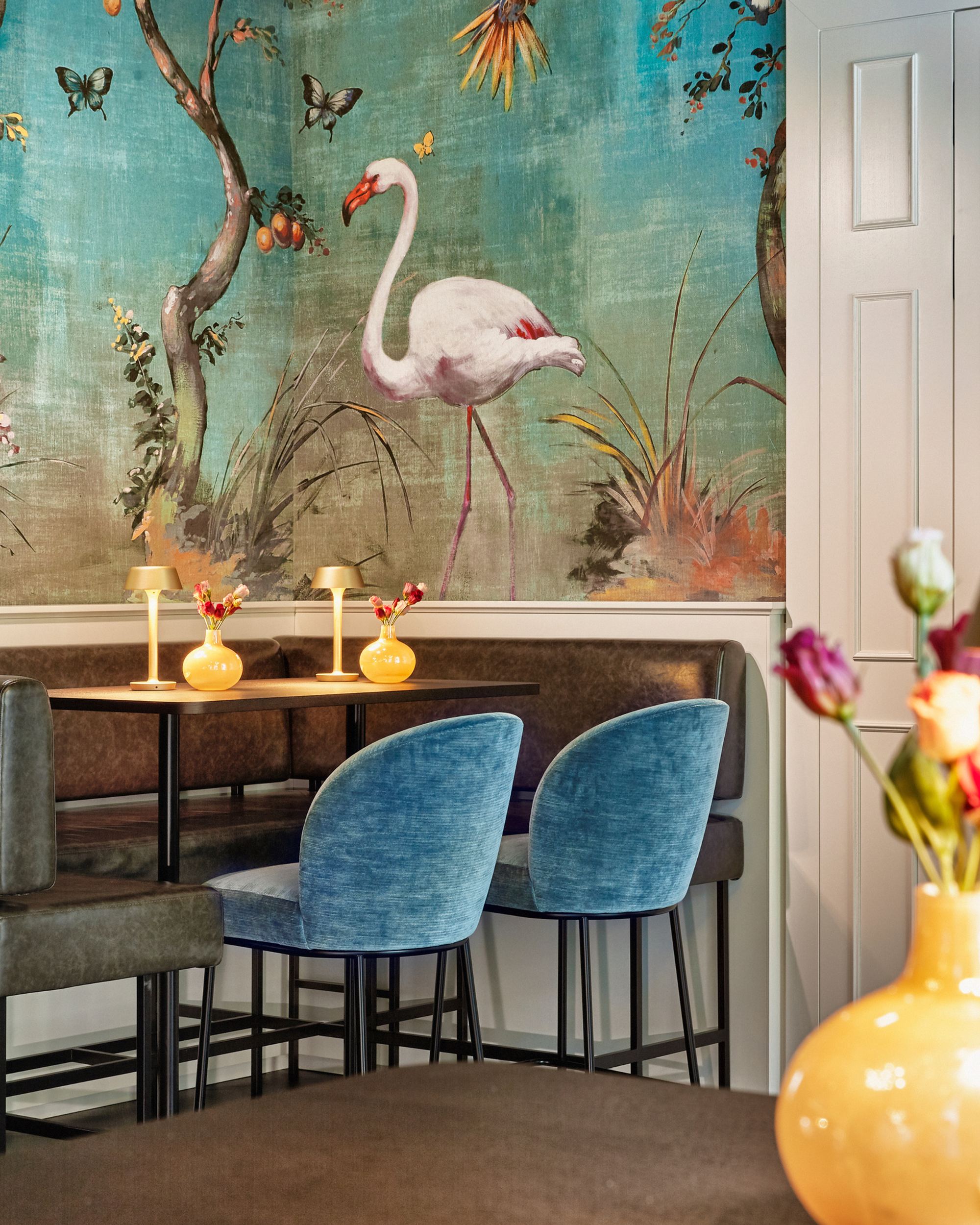 Gemütliche Sitzecke mit dunkelblauen Stühlen und brauner Sitzbank vor einer Wand mit farbenfrohem Flamingo-Tapetenmotiv, dekoriert mit Blumen und sanfter Beleuchtung für eine entspannte Atmosphäre.