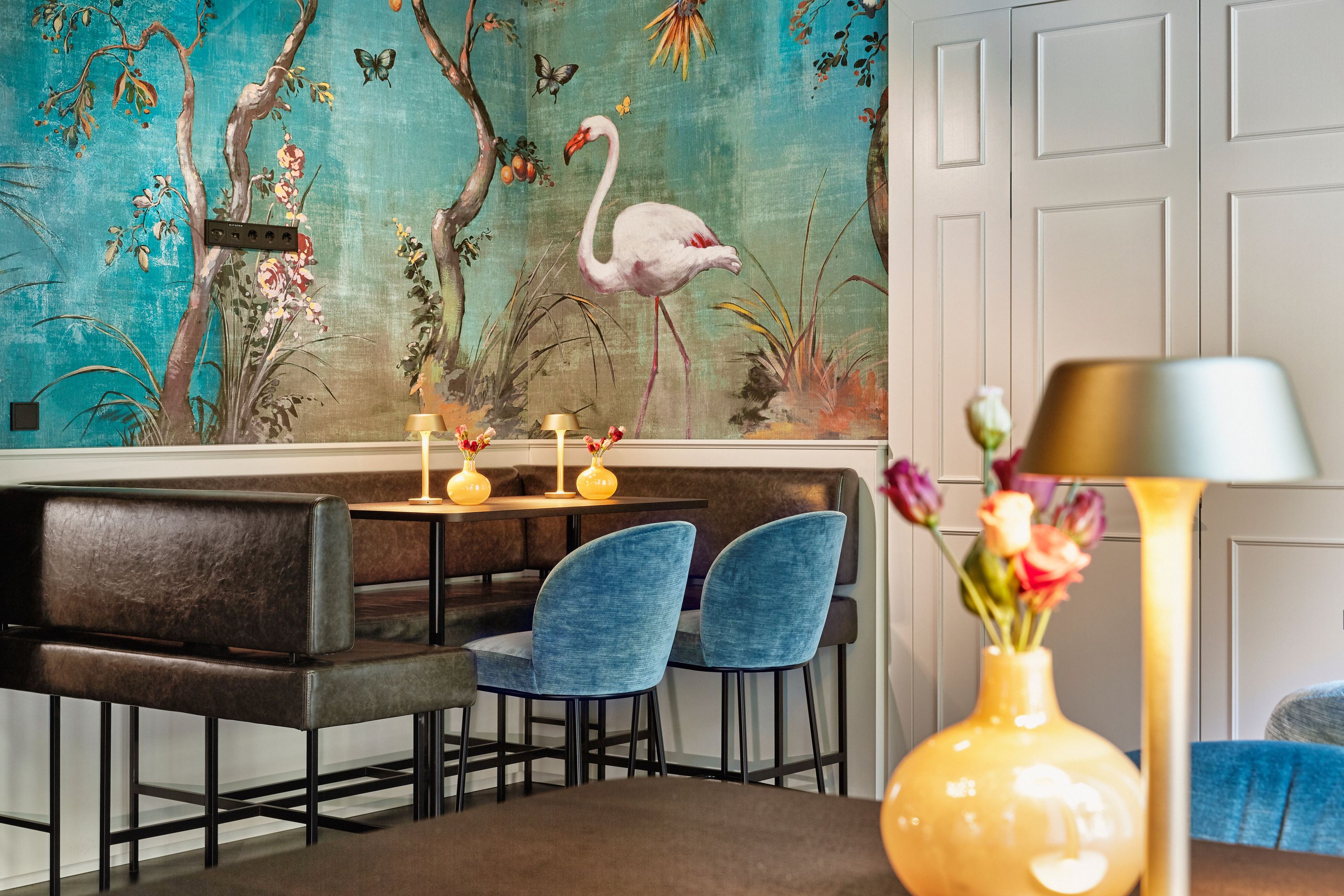 Gemütliche Sitzecke mit dunkelblauen Stühlen und brauner Sitzbank vor einer Wand mit farbenfrohem Flamingo-Tapetenmotiv, dekoriert mit Blumen und sanfter Beleuchtung für eine entspannte Atmosphäre. Gemütliche Sitzecke mit dunkelblauen Stühlen und brauner Sitzbank vor einer Wand mit farbenfrohem Flamingo-Tapetenmotiv, dekoriert mit Blumen und sanfter Beleuchtung für eine entspannte Atmosphäre.