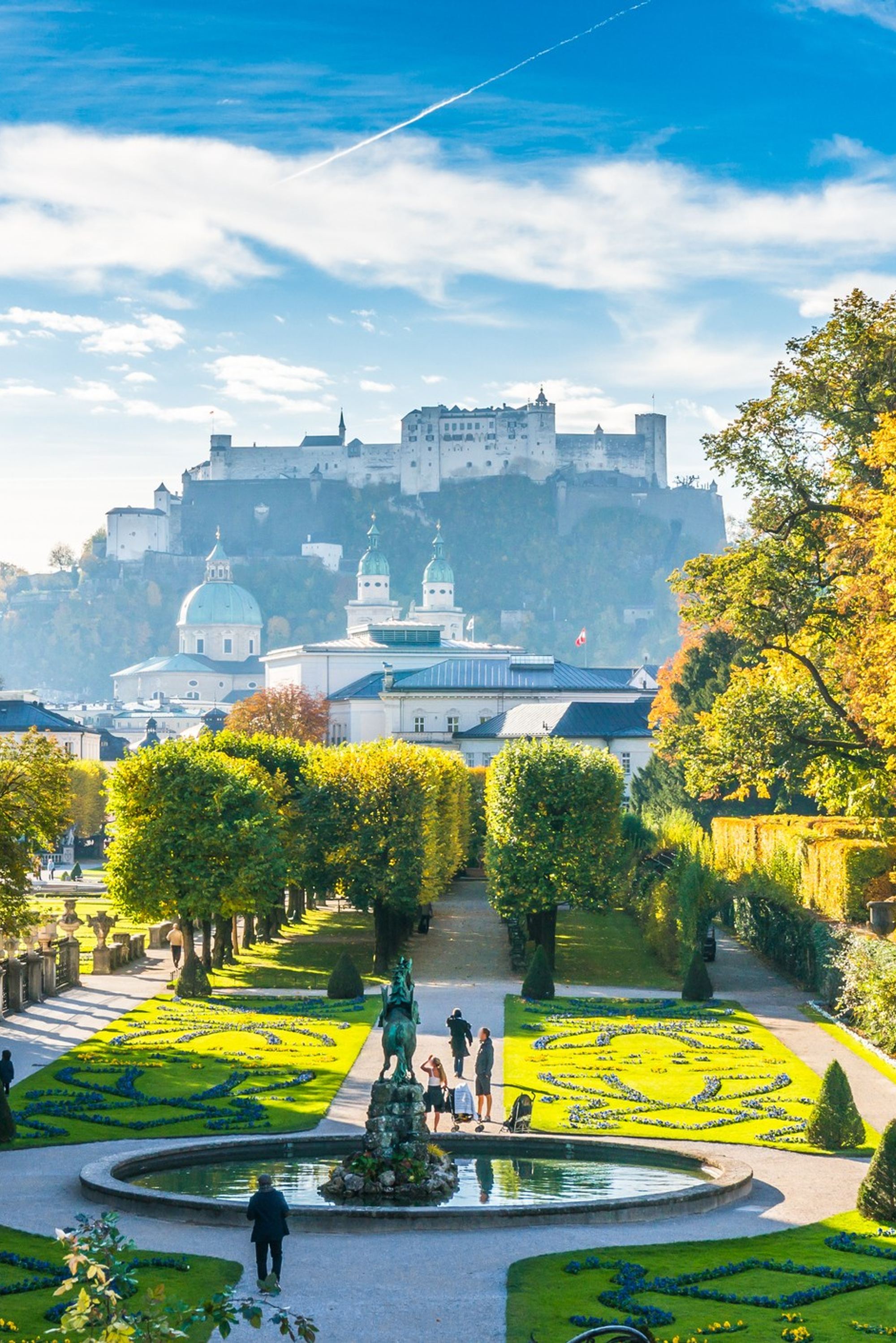 Blick auf einen gepflegten Barockgarten mit kunstvoll geschnittenen Hecken und Springbrunnen, umgeben von historischen Gebäuden, mit der Festung Hohensalzburg im Hintergrund unter blauem Himmel.