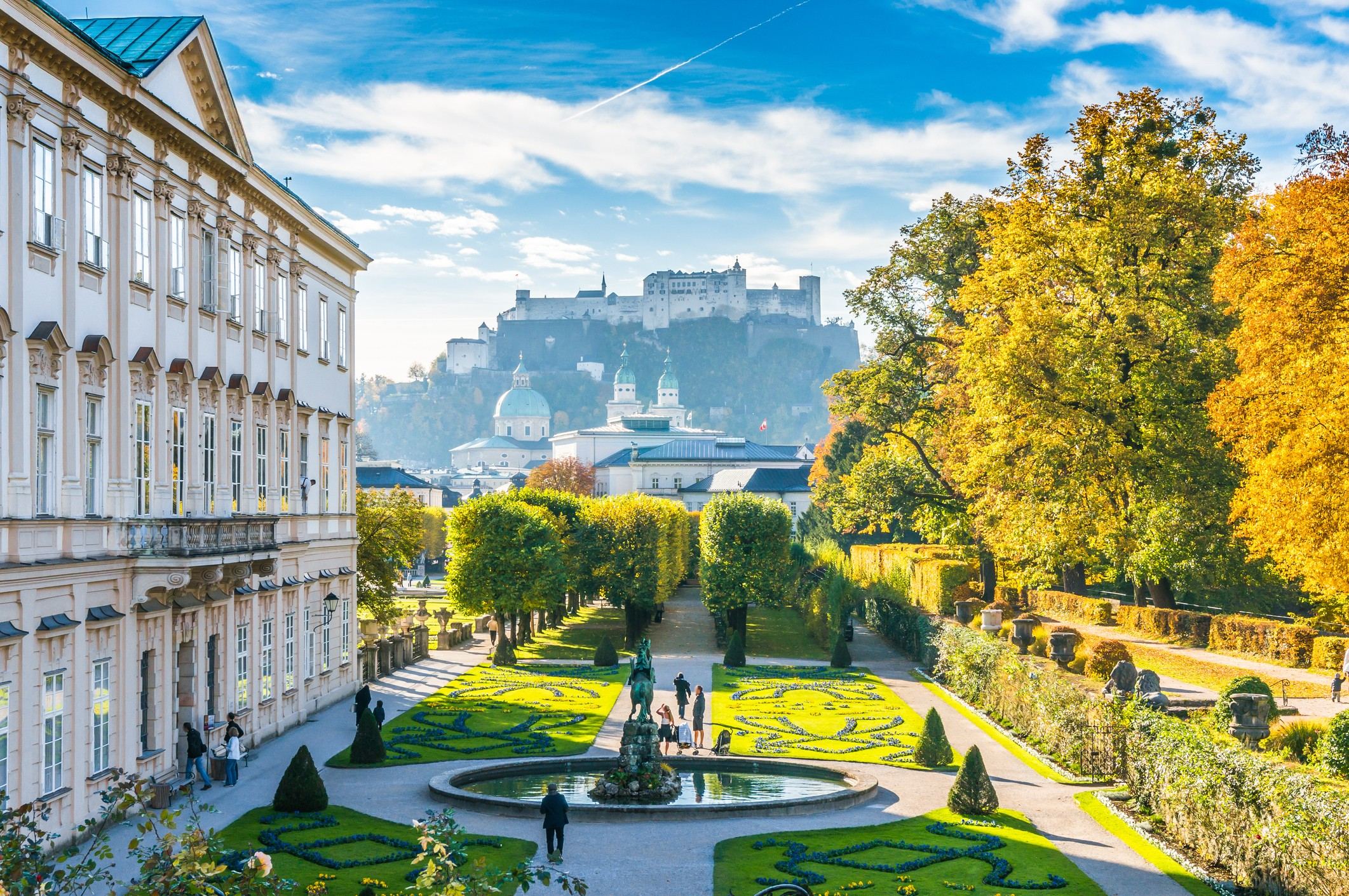 Blick auf einen gepflegten Barockgarten mit kunstvoll geschnittenen Hecken und Springbrunnen, umgeben von historischen Gebäuden, mit der Festung Hohensalzburg im Hintergrund unter blauem Himmel. Blick auf einen gepflegten Barockgarten mit kunstvoll geschnittenen Hecken und Springbrunnen, umgeben von historischen Gebäuden, mit der Festung Hohensalzburg im Hintergrund unter blauem Himmel.