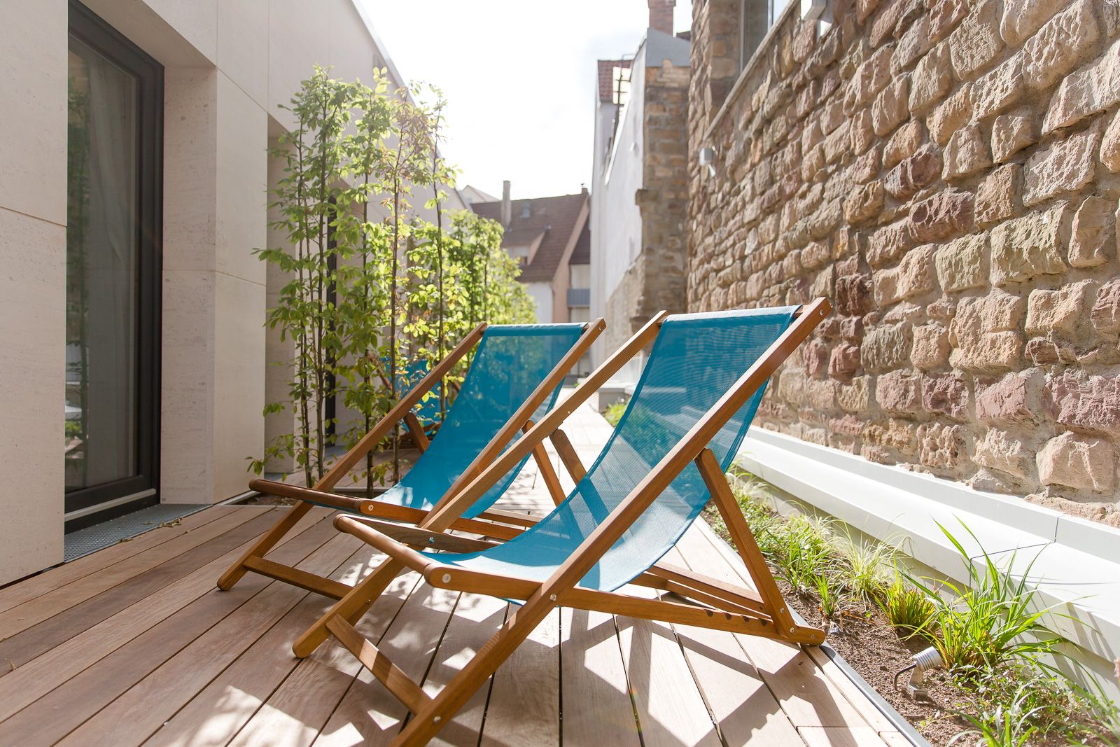Zwei blaue Liegestühle auf einer sonnigen Holzterrasse neben einer Backsteinwand, umgeben von Pflanzen, laden zum Entspannen ein.