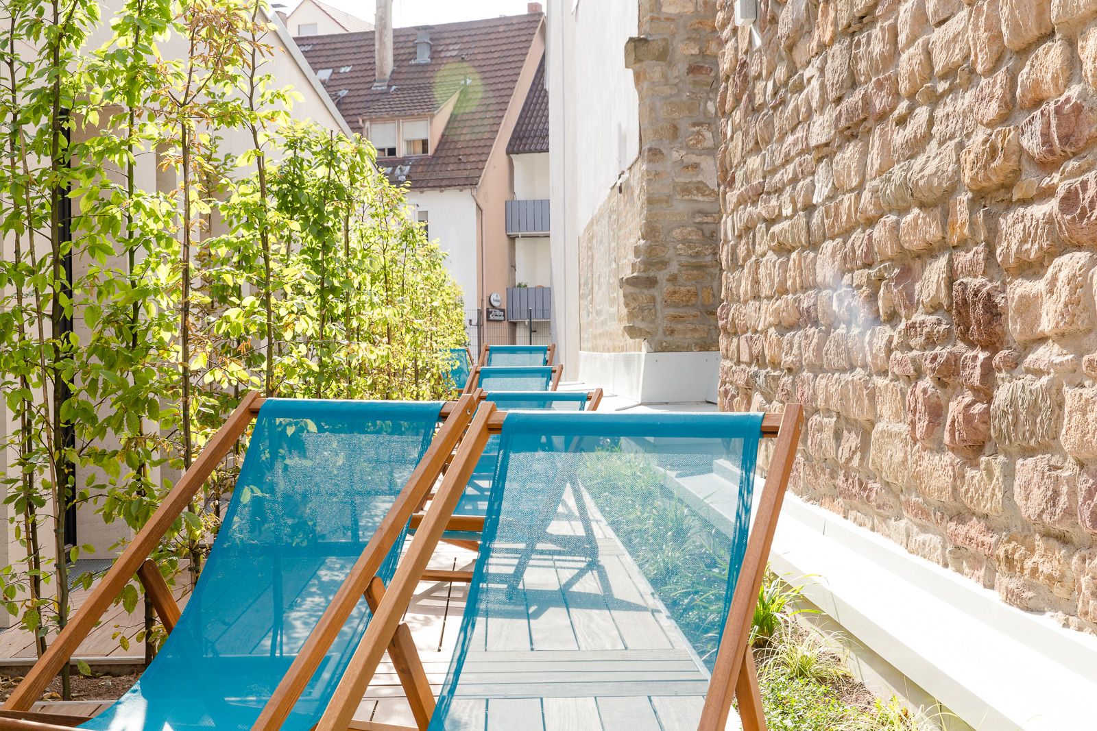 Mehrere türkisfarbene Liegestühle stehen auf einem sonnigen, schmalen Innenhof mit Natursteinmauer und grünen Kletterpflanzen, ideal zum Entspannen im Freien.