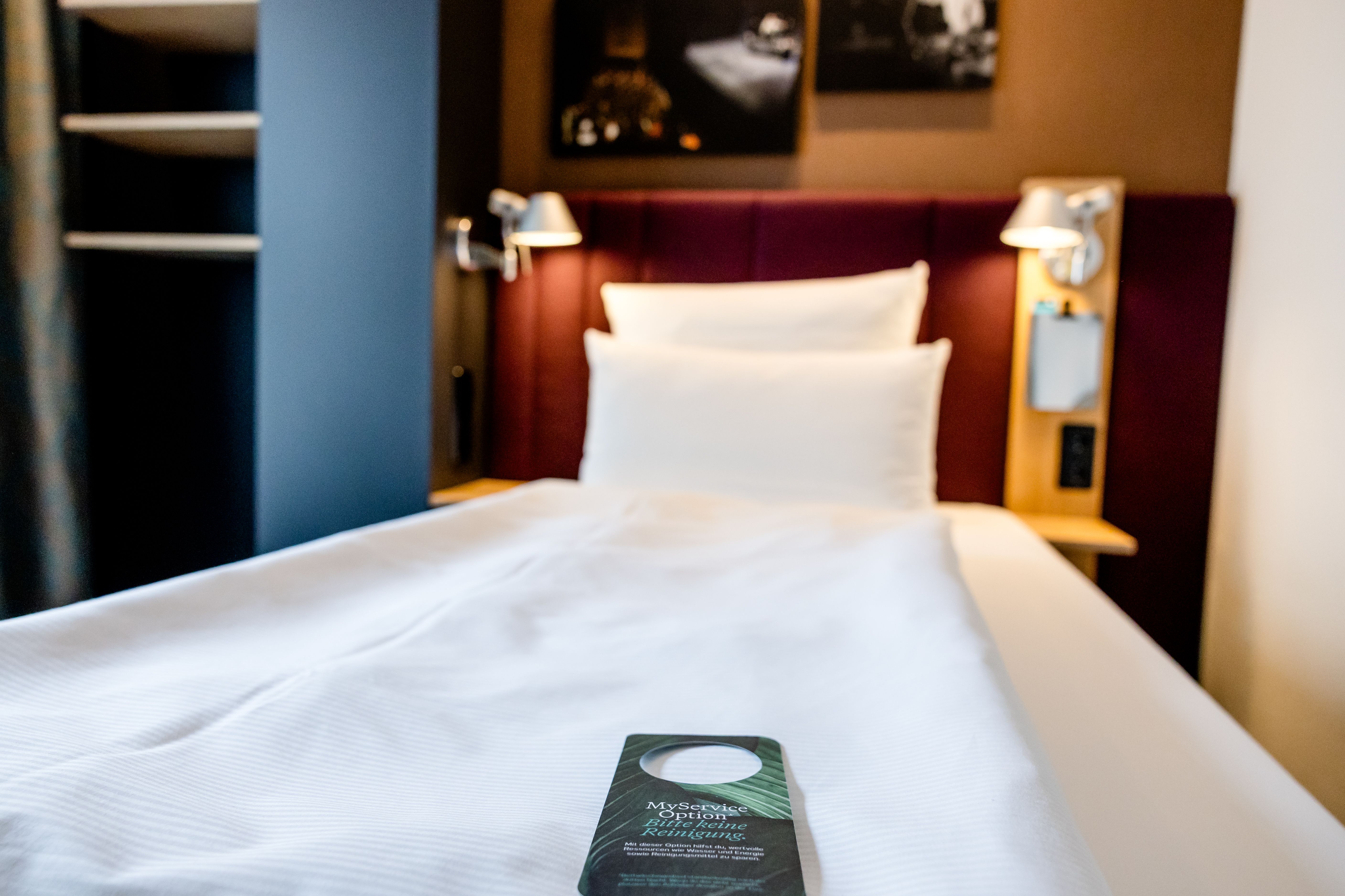 Schmales Einzelbett mit weißer Bettwäsche, zwei Kissen und rotem Polsterkopfteil in einem modernen Zimmer, mit Leselampen und einem Hinweiszettel auf der Bettdecke.
