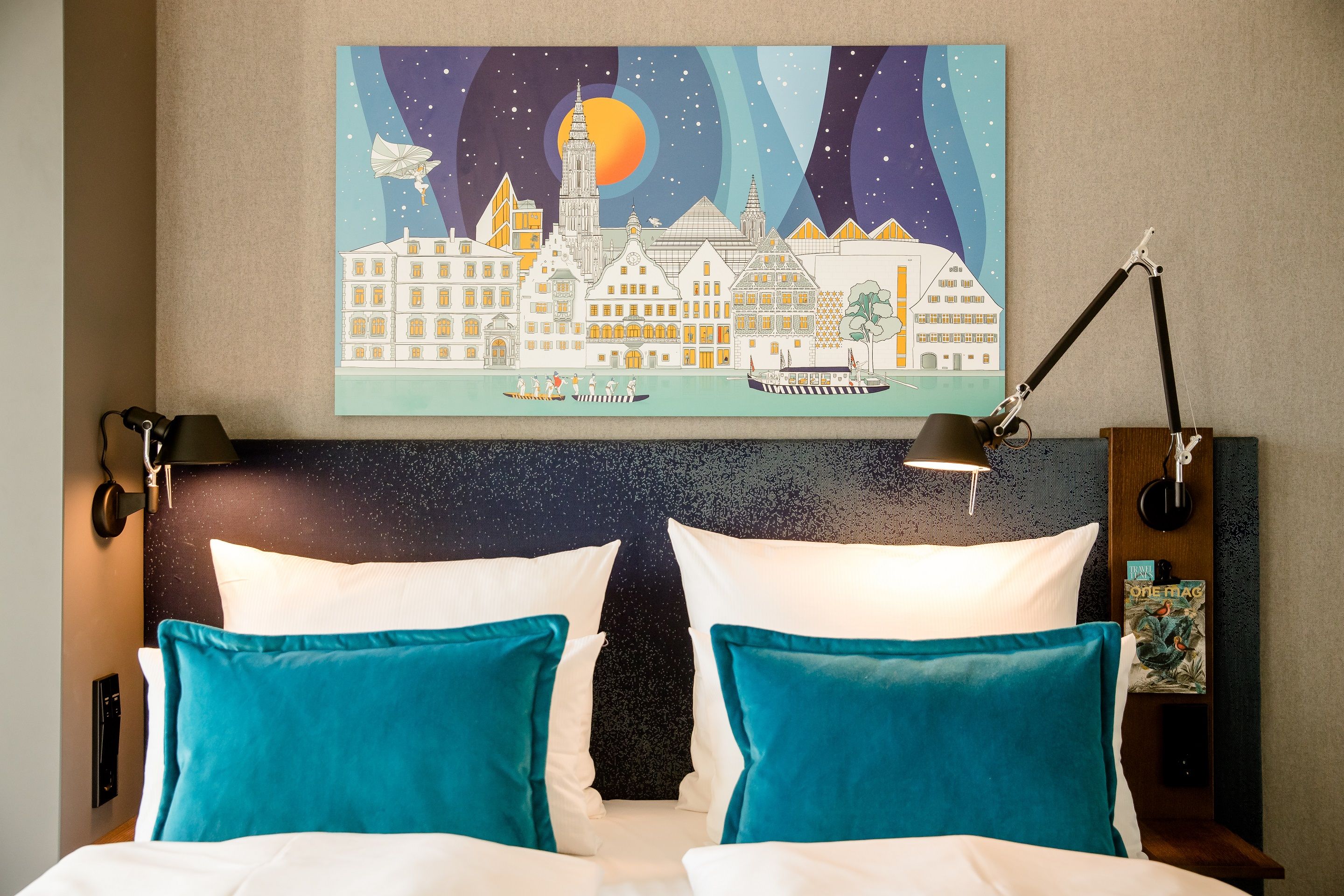 Moderner Hotelzimmer-Ausschnitt mit komfortablem Doppelbett, hellen weißen Kissen und zwei türkisblauen Dekokissen, beleuchtet von flexiblen Wandlampen. Über dem Bett hängt ein farbenfrohes Bild einer Stadtansicht bei Nacht.