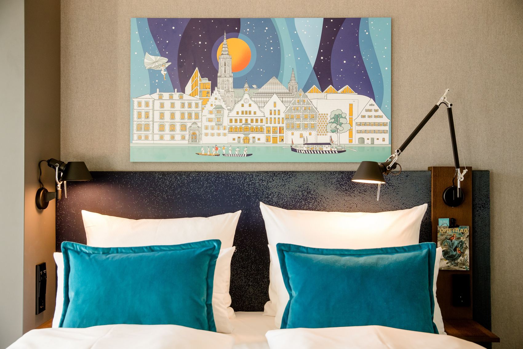 Moderner Hotelzimmer-Ausschnitt mit komfortablem Doppelbett, hellen weißen Kissen und zwei türkisblauen Dekokissen, beleuchtet von flexiblen Wandlampen. Über dem Bett hängt ein farbenfrohes Bild einer Stadtansicht bei Nacht.