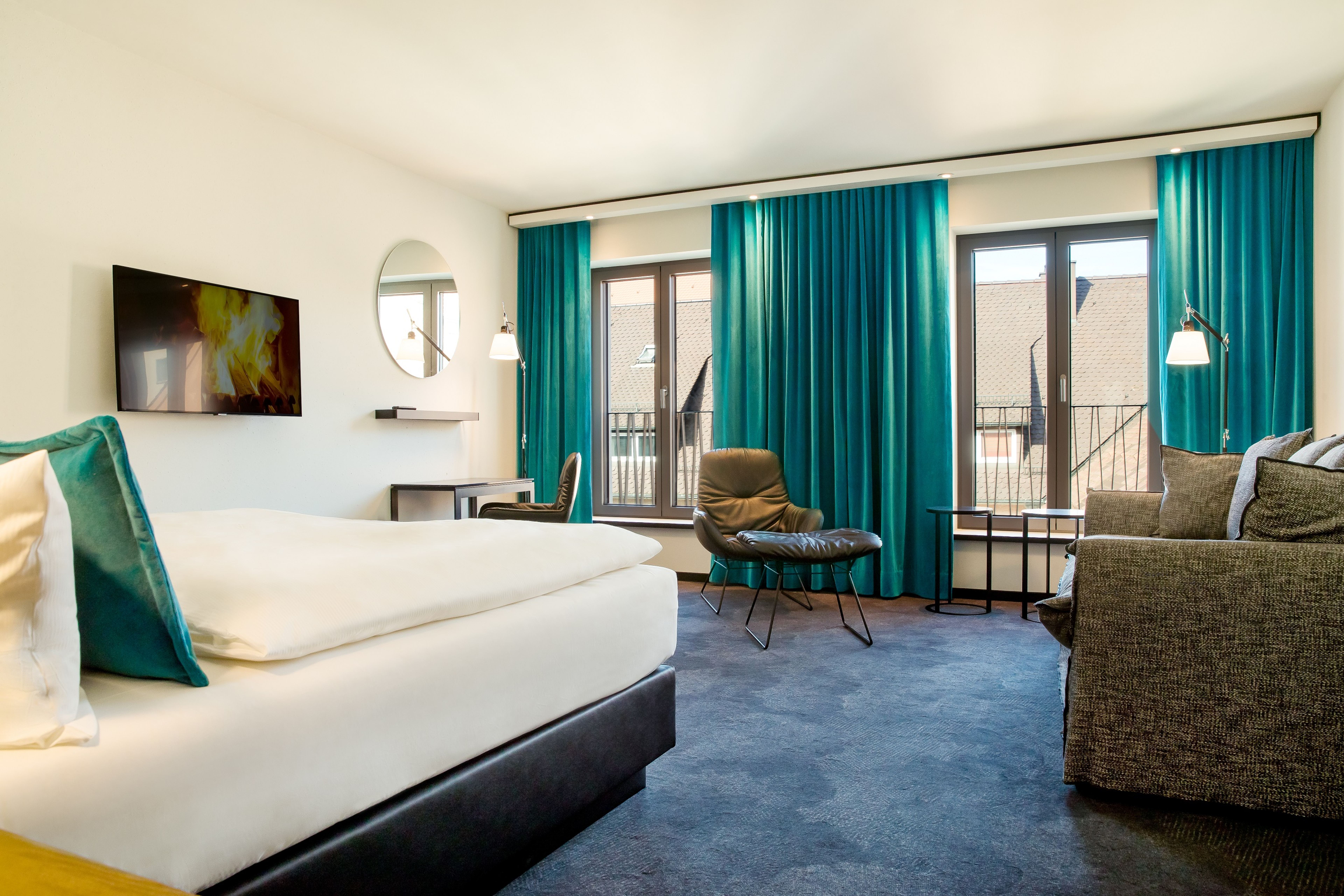 Hell eingerichtetes Hotelzimmer mit großem Bett, blauem Teppich, türkisfarbenen Vorhängen sowie Sitzbereich mit Sessel und Sofa vor bodentiefen Fenstern, die viel Tageslicht ins Zimmer lassen. Hell eingerichtetes Hotelzimmer mit großem Bett, blauem Teppich, türkisfarbenen Vorhängen sowie Sitzbereich mit Sessel und Sofa vor bodentiefen Fenstern, die viel Tageslicht ins Zimmer lassen.