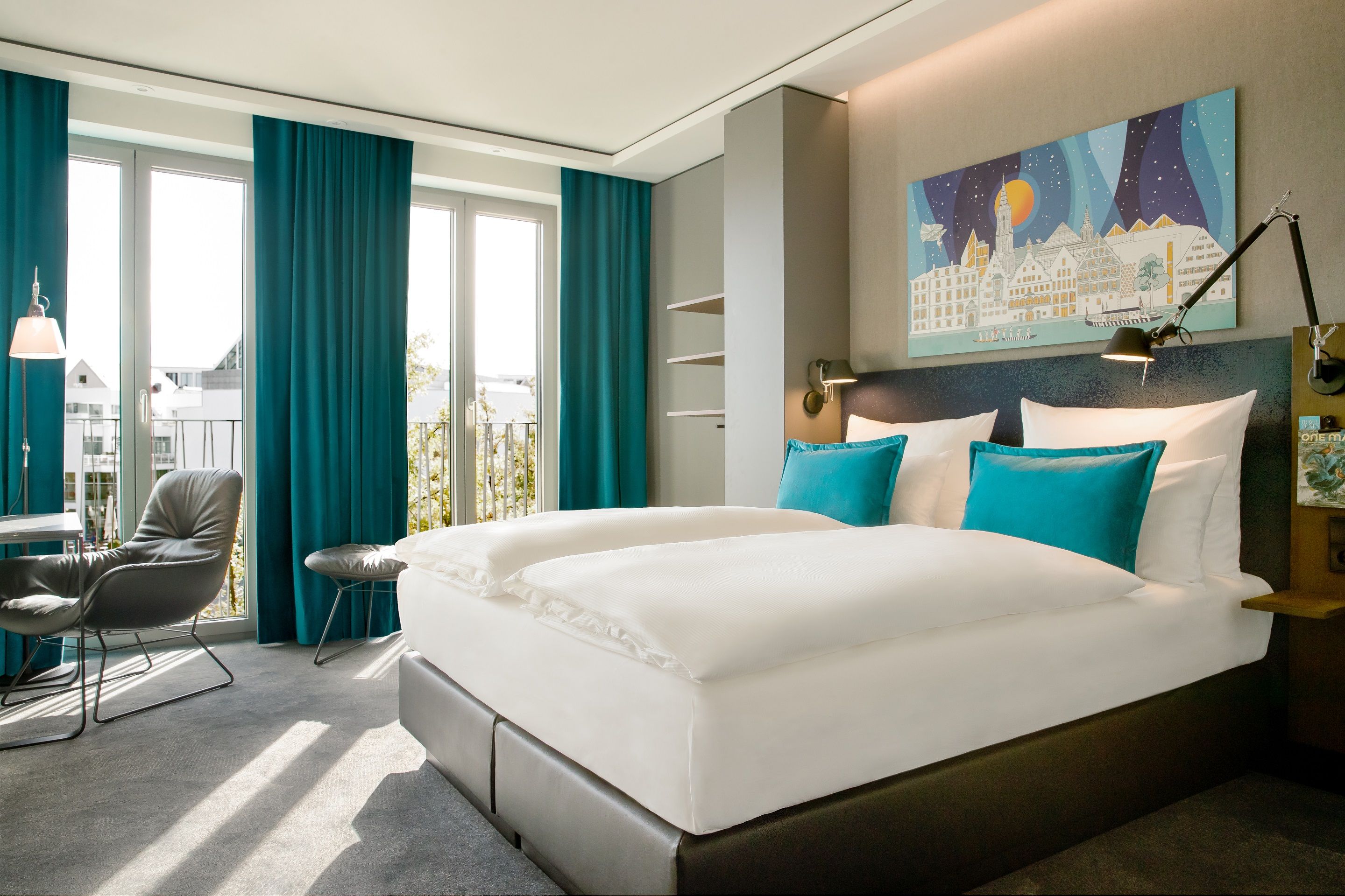 Helles Hotelzimmer mit großem Doppelbett, türkisfarbenen Kissen und Vorhängen, Sitzbereich mit Stuhl und Tisch sowie Panoramafenstern, die viel Tageslicht und Ausblick bieten, modernes und gemütliches Ambiente.