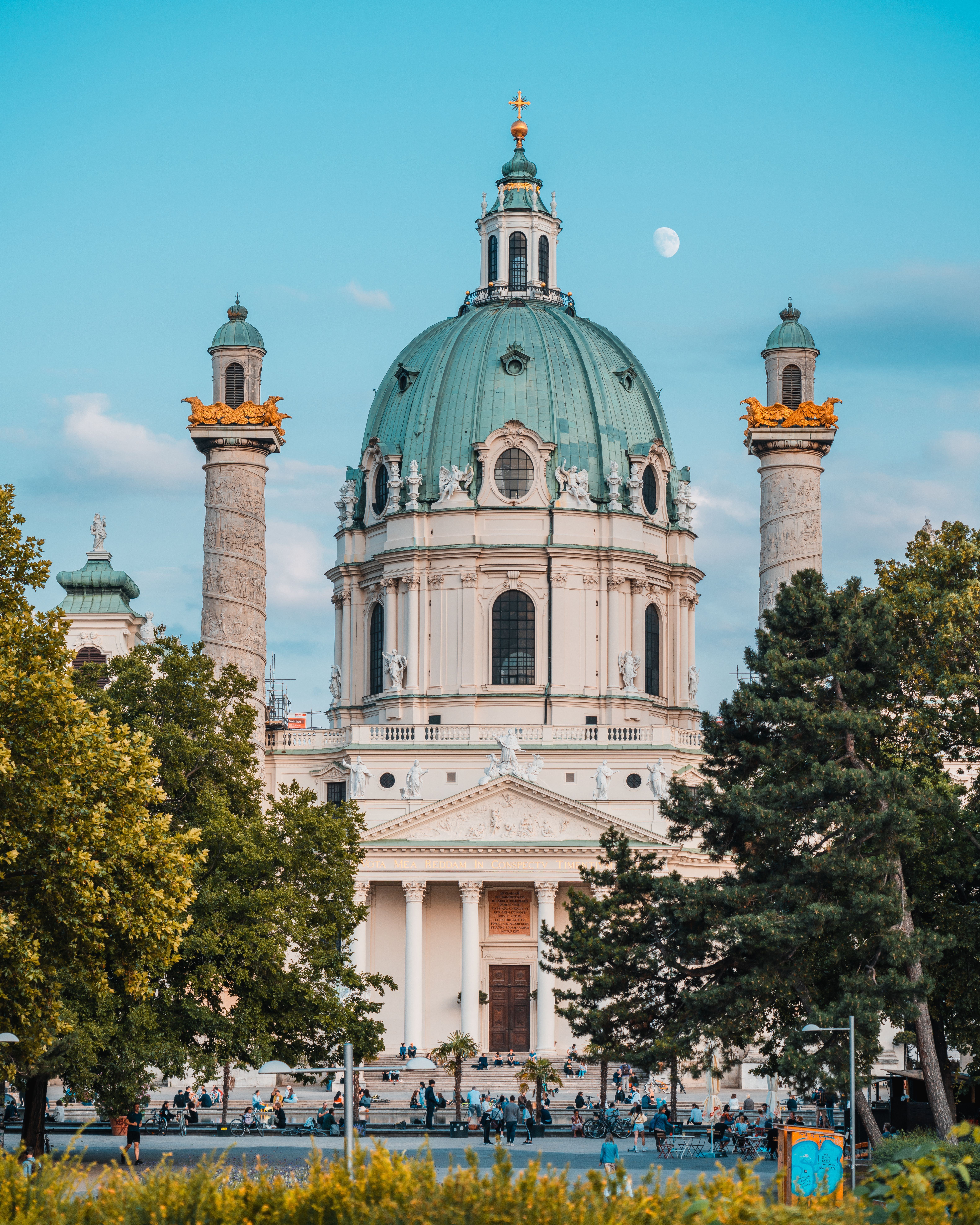 Barocke Kirche mit großer grüner Kuppel und zwei verzierenden Säulen, umgeben von Bäumen, bei klarem Himmel und sichtbarem Mond, mit Personen auf dem Vorplatz.