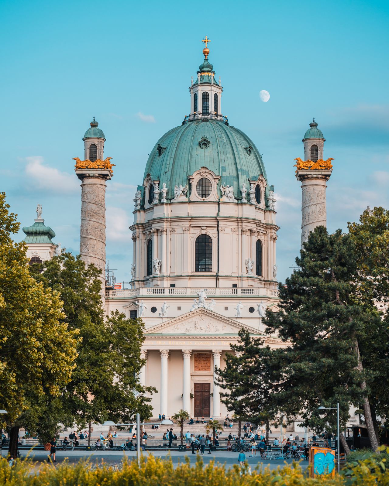 Barocke Kirche mit großer grüner Kuppel und zwei verzierenden Säulen, umgeben von Bäumen, bei klarem Himmel und sichtbarem Mond, mit Personen auf dem Vorplatz.