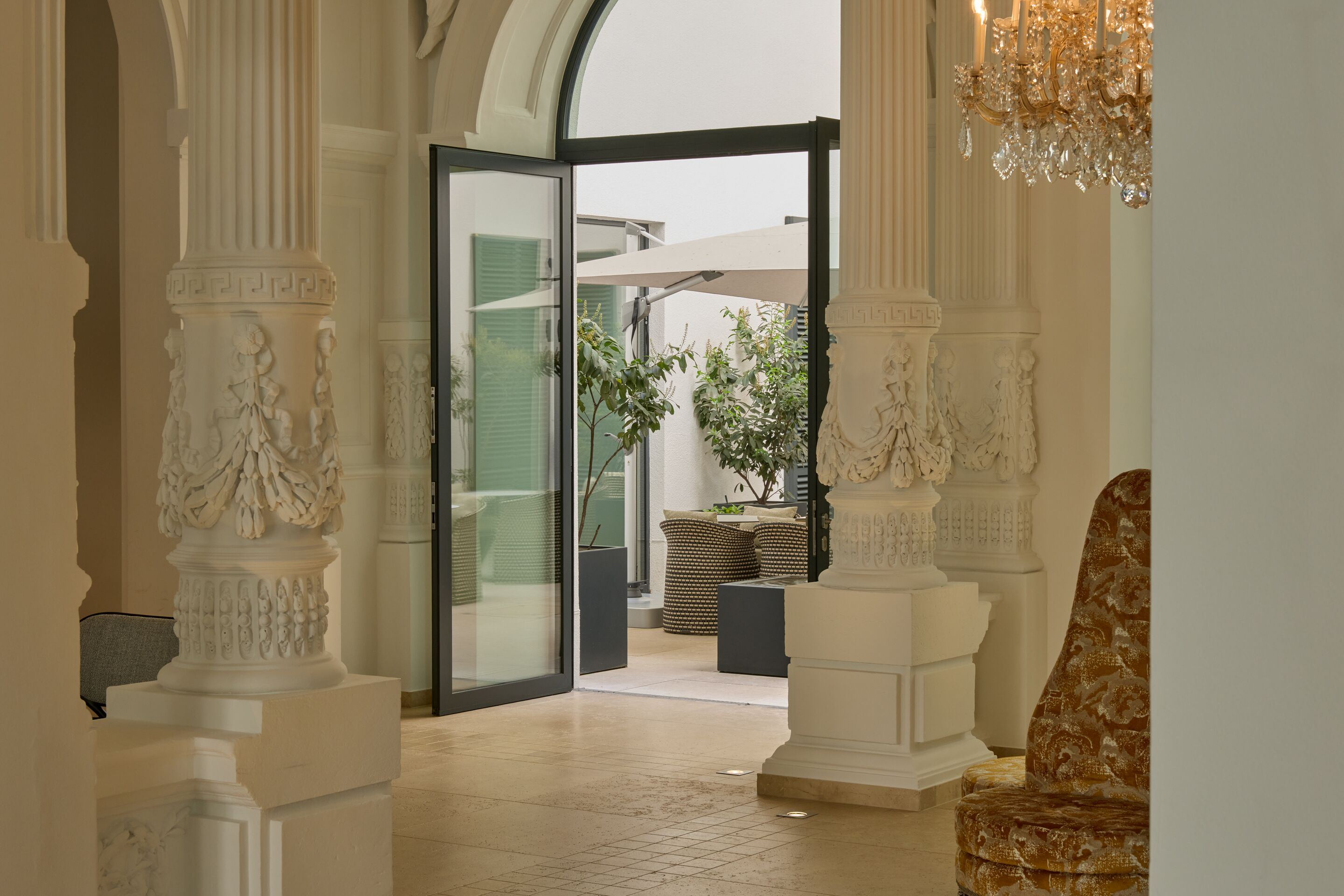 Heller, eleganter Innenraum mit prächtigen, verzierten Säulen und offenem Eingang zu einem Außenbereich mit Pflanzen und Sitzgelegenheiten unter Sonnenschirmen. Heller, eleganter Innenraum mit prächtigen, verzierten Säulen und offenem Eingang zu einem Außenbereich mit Pflanzen und Sitzgelegenheiten unter Sonnenschirmen.