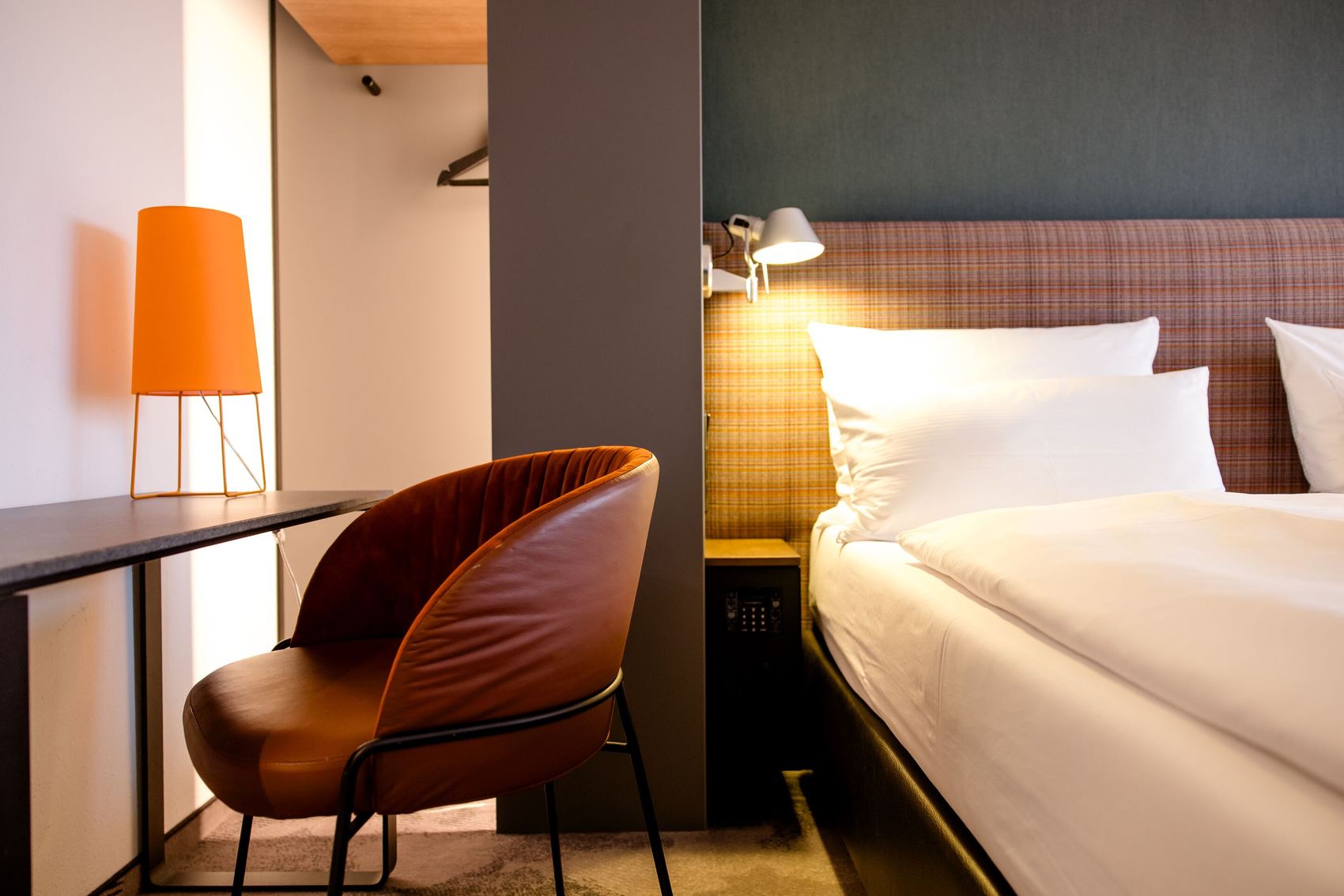 Hotel Motel One Wien-Prater Moderner Hotelzimmerbereich mit gemütlichem Bett, warmen Holztönen, einem bequemen Stuhl und dezent beleuchtetem Arbeitsplatz für produktives Arbeiten oder Entspannen.