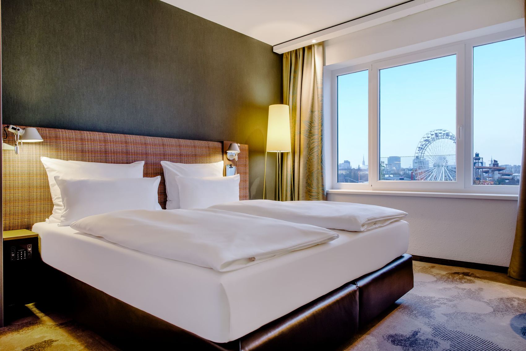Hotel Motel One Wien-Prater Hell eingerichtetes Hotelzimmer mit großem gemütlichem Doppelbett, warmem Licht und Fenster mit Blick auf ein Riesenrad und die Stadt. Atmosphäre lädt zum Entspannen nach einem ereignisreichen Tag ein.