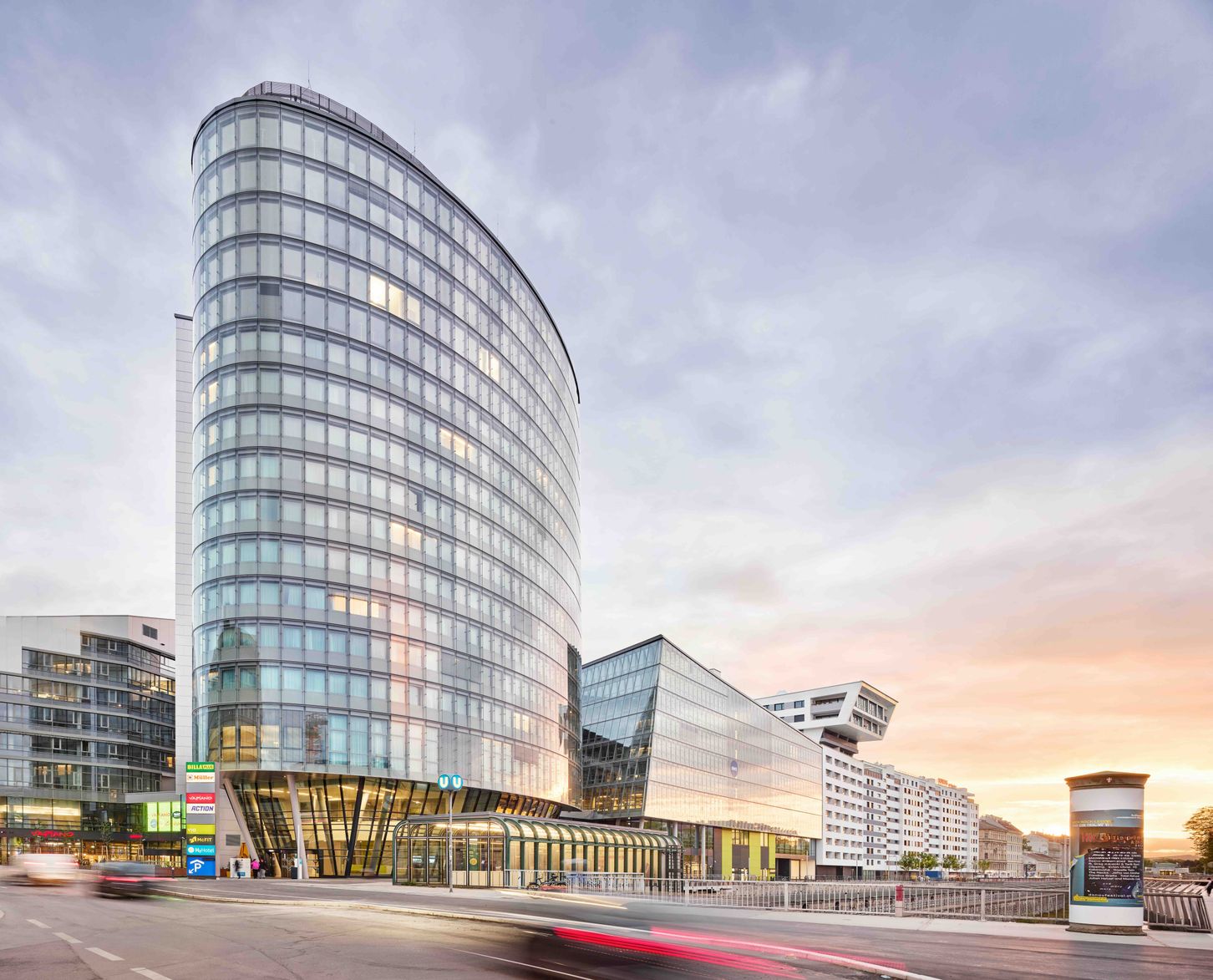 Modernes, elegantes Hotelgebäude mit geschwungener Glasfassade, das städtisches, urbanes Flair vermittelt. Ideal für Geschäftsreisende und Stadtentdecker, zentrale Lage mit guter Anbindung bei Sonnenuntergang.