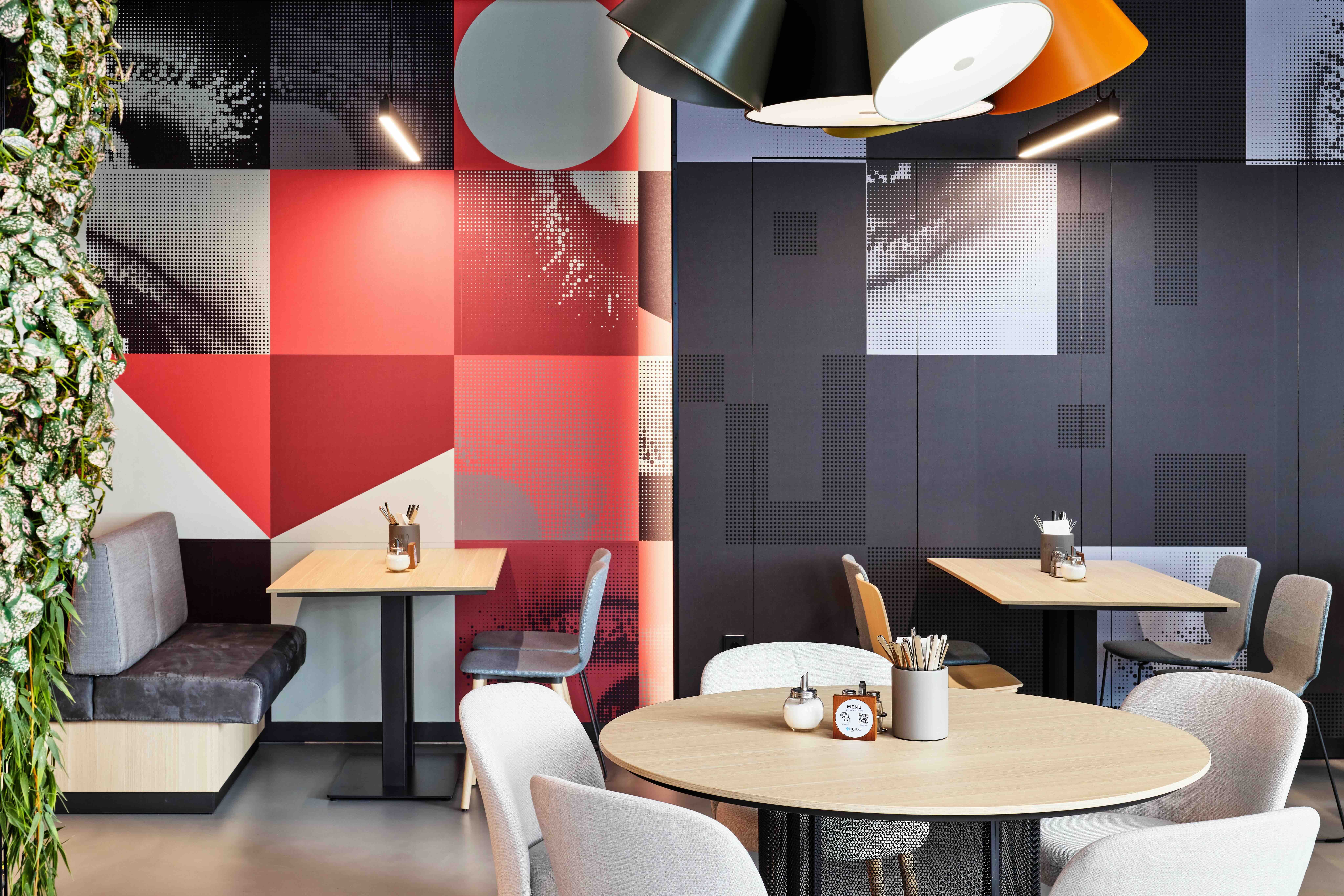 Moderner Sitzbereich mit runden und eckigen Tischen, bequemen Stühlen und stilvoller Wandgestaltung in Rot-, Schwarz- und Grautönen, beleuchtet von großer Deckenlampe, vermittelt ein entspanntes, urbanes Ambiente zum Essen oder Arbeiten.
