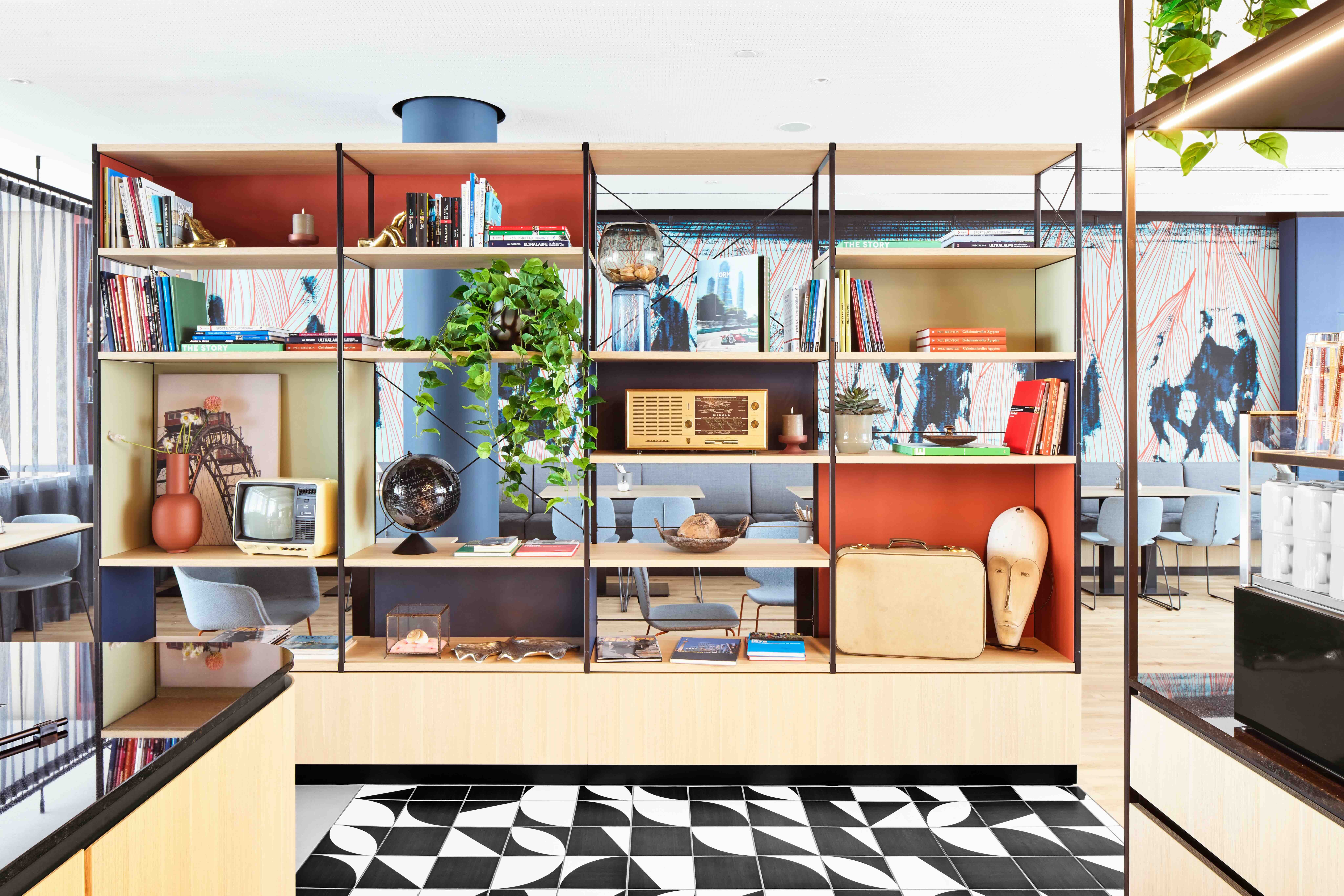 Offene Regalwand mit Büchern, Pflanzen, historischen Radios und Dekorationsobjekten, die eine moderne, gemütliche Atmosphäre im Hotel-Loungebereich schafft.