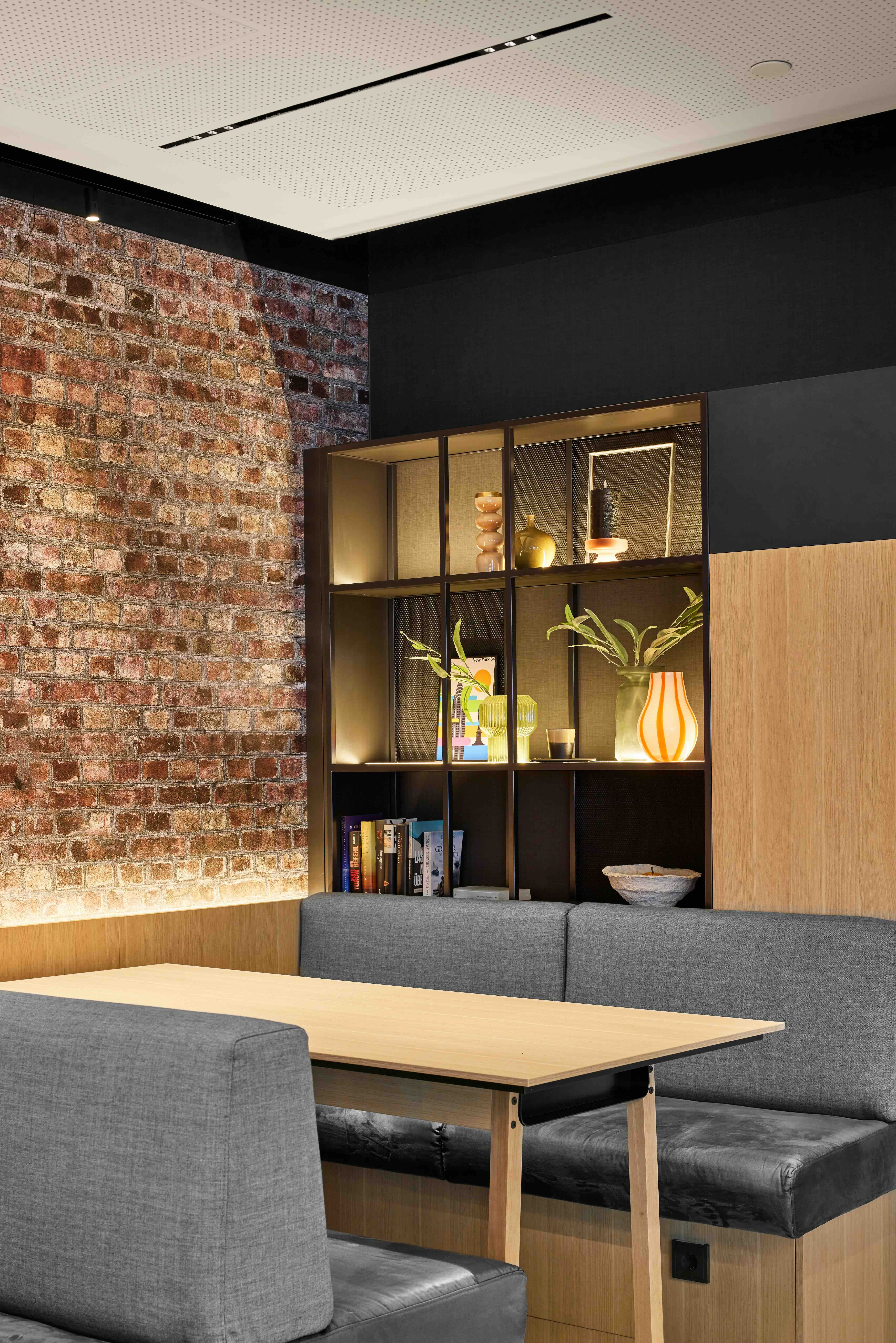 Gemütliche Sitzecke mit grauen Polstersesseln und Holztisch vor einer beleuchteten Regalwand. Backsteinwand und moderne, warme Beleuchtung schaffen eine einladende Atmosphäre. Ideal zum Entspannen oder für Gespräche.