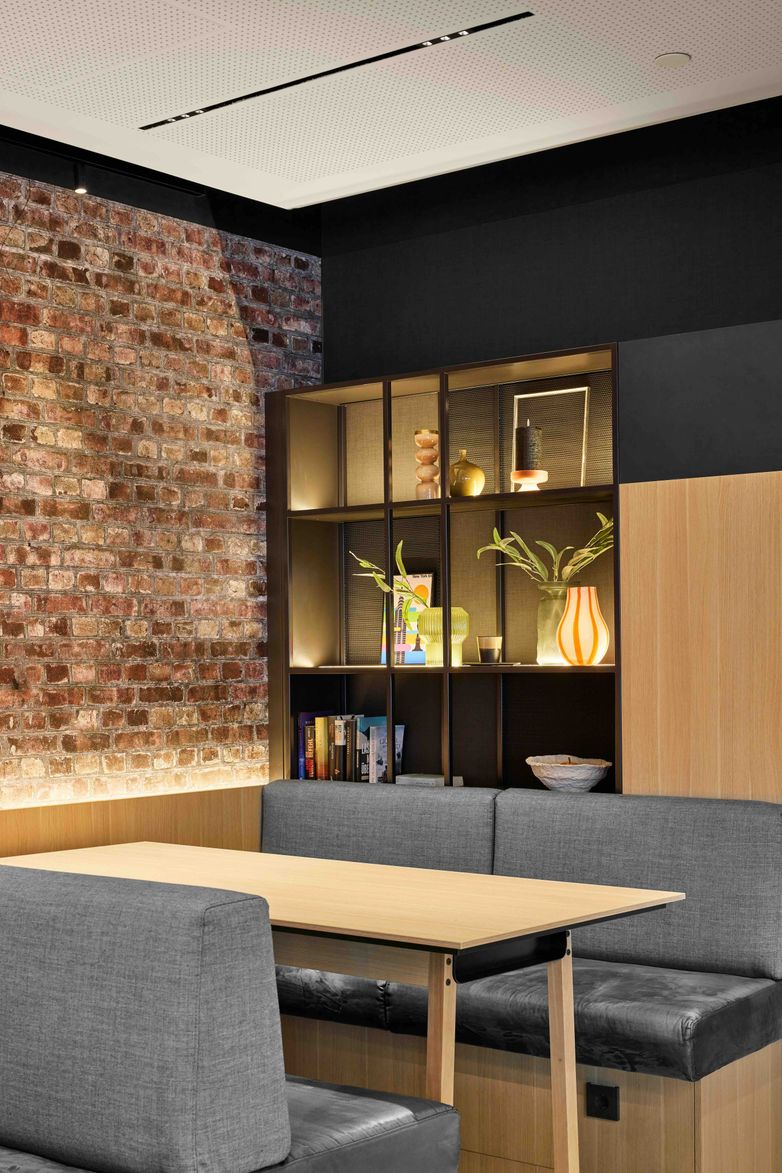 Gemütliche Sitzecke mit grauen Polstersesseln und Holztisch vor einer beleuchteten Regalwand. Backsteinwand und moderne, warme Beleuchtung schaffen eine einladende Atmosphäre. Ideal zum Entspannen oder für Gespräche.