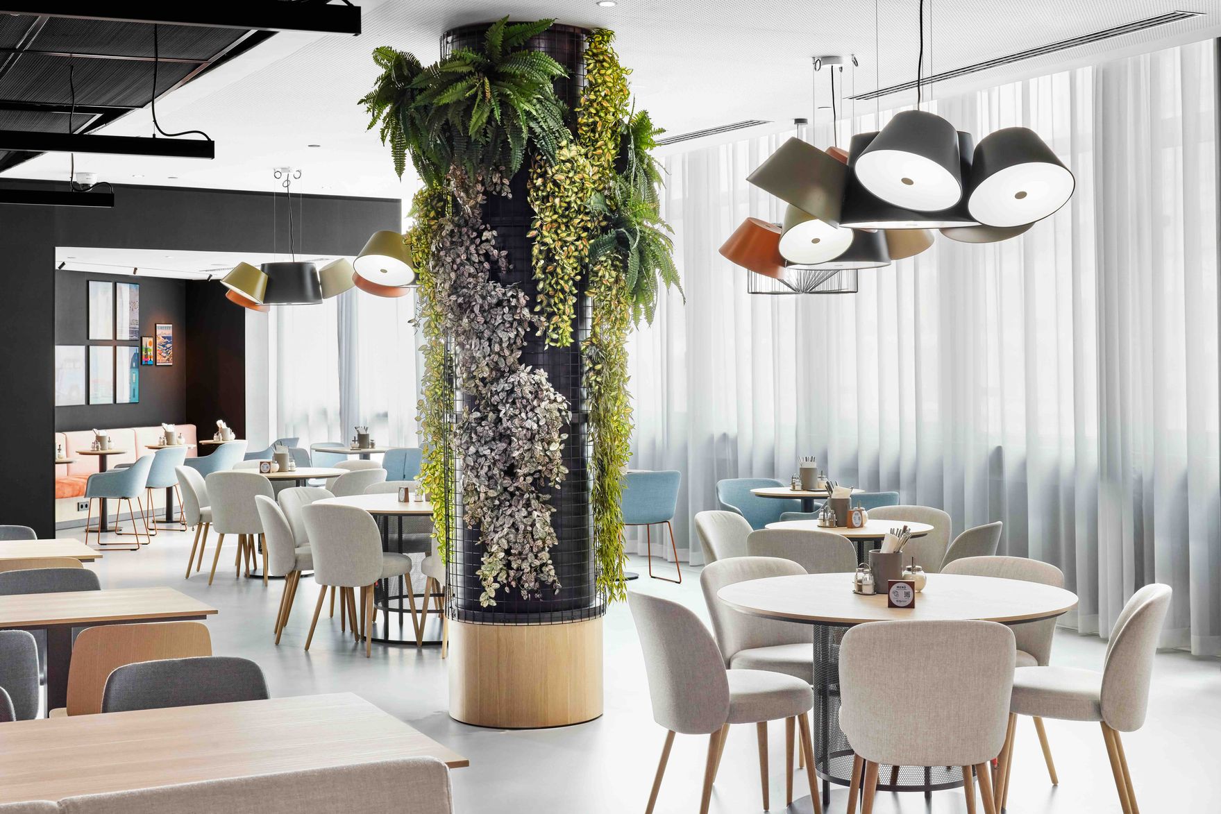 Moderner, heller Restaurantbereich mit runden Tischen, komfortablen Stühlen und einer Säule, die mit grünen und weißen Pflanzen dekoriert ist, sowie stilvollen Deckenlampen, die ein einladendes Ambiente schaffen. Große Fenster lassen viel Tageslicht herein.