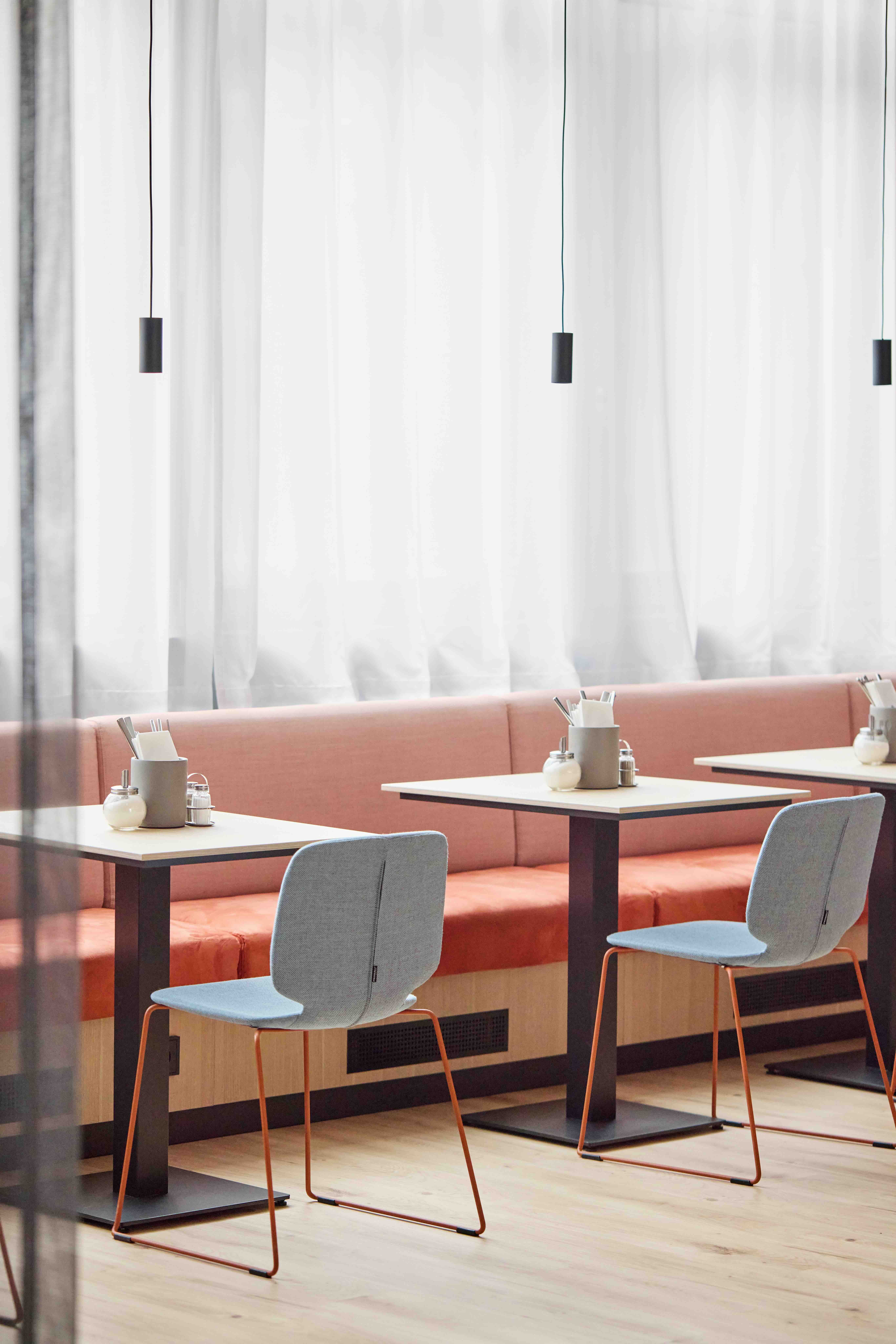 Moderner Essbereich mit hellen Stühlen, rosafarbenem Sofa und kleinen Tischen vor lichtdurchlässigen Vorhängen, schafft eine einladende und ruhige Atmosphäre zum Speisen.