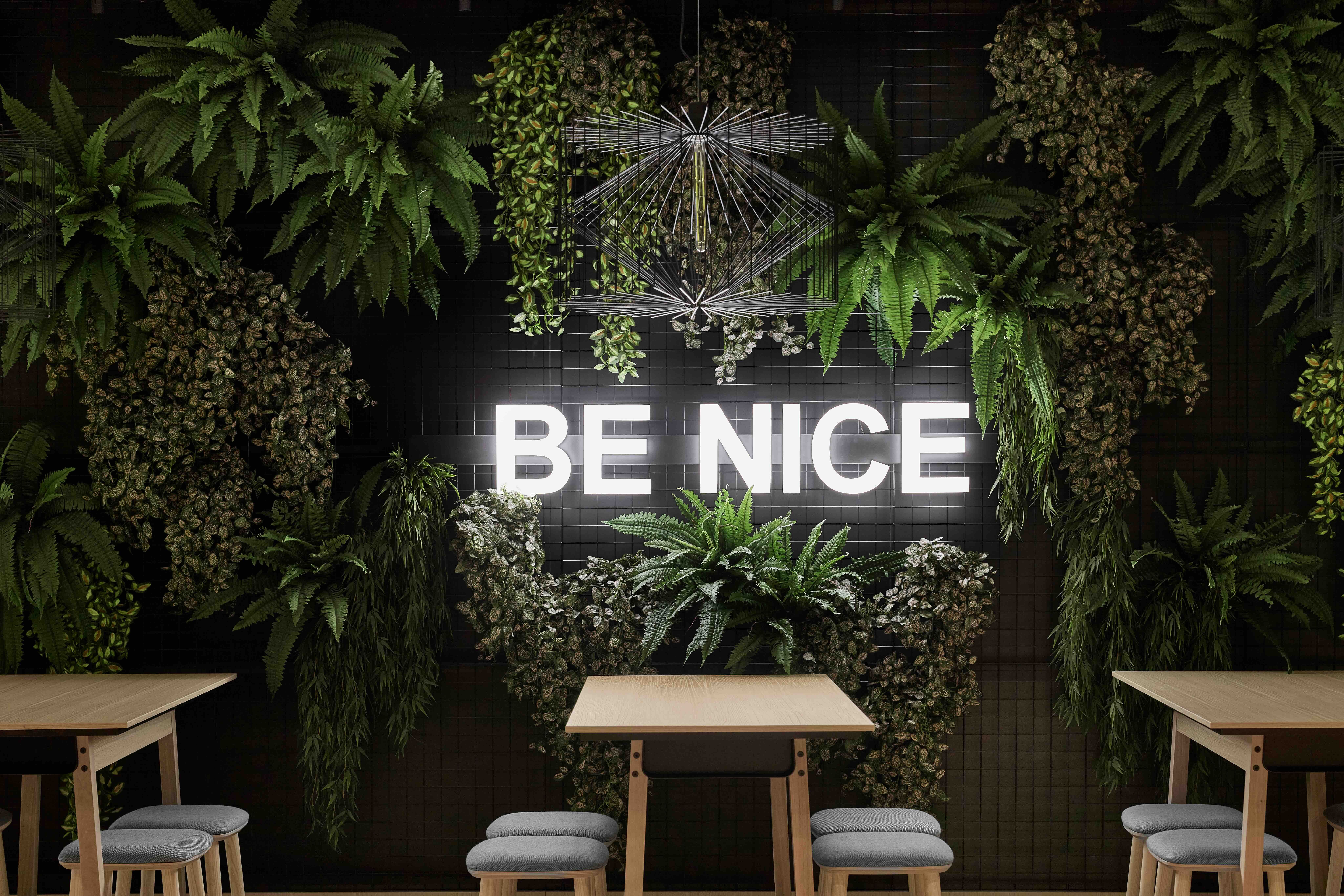 Leuchtendes Schild mit dem Text „BE NICE“ an einer Wand aus grünen Pflanzen, darüber moderne Beleuchtung. Darunter einfache Holztische mit grauen Sitzhockern in einem einladenden, naturverbundenen Ambiente.