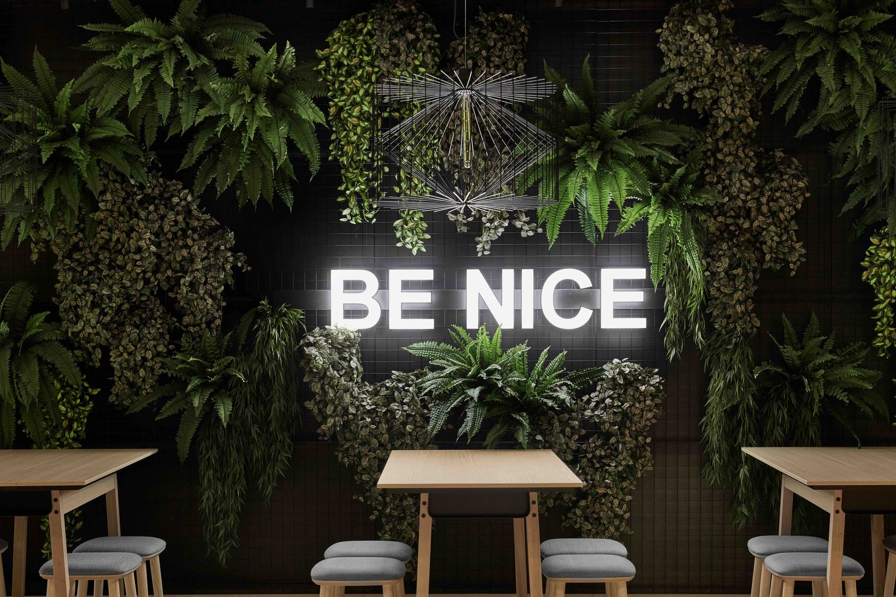 Leuchtendes Schild mit dem Text „BE NICE“ an einer Wand aus grünen Pflanzen, darüber moderne Beleuchtung. Darunter einfache Holztische mit grauen Sitzhockern in einem einladenden, naturverbundenen Ambiente.