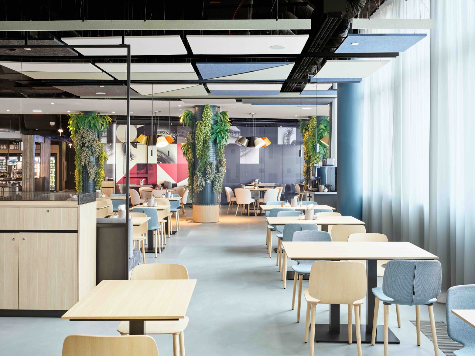 Moderner, heller Restaurantbereich mit hellen Holztischen und Stühlen, dezenten Pastellfarben und großen Pflanzen. Großzügige Fensterfront sorgt für viel Tageslicht und eine freundliche Atmosphäre zum entspannten Essen oder Arbeiten.