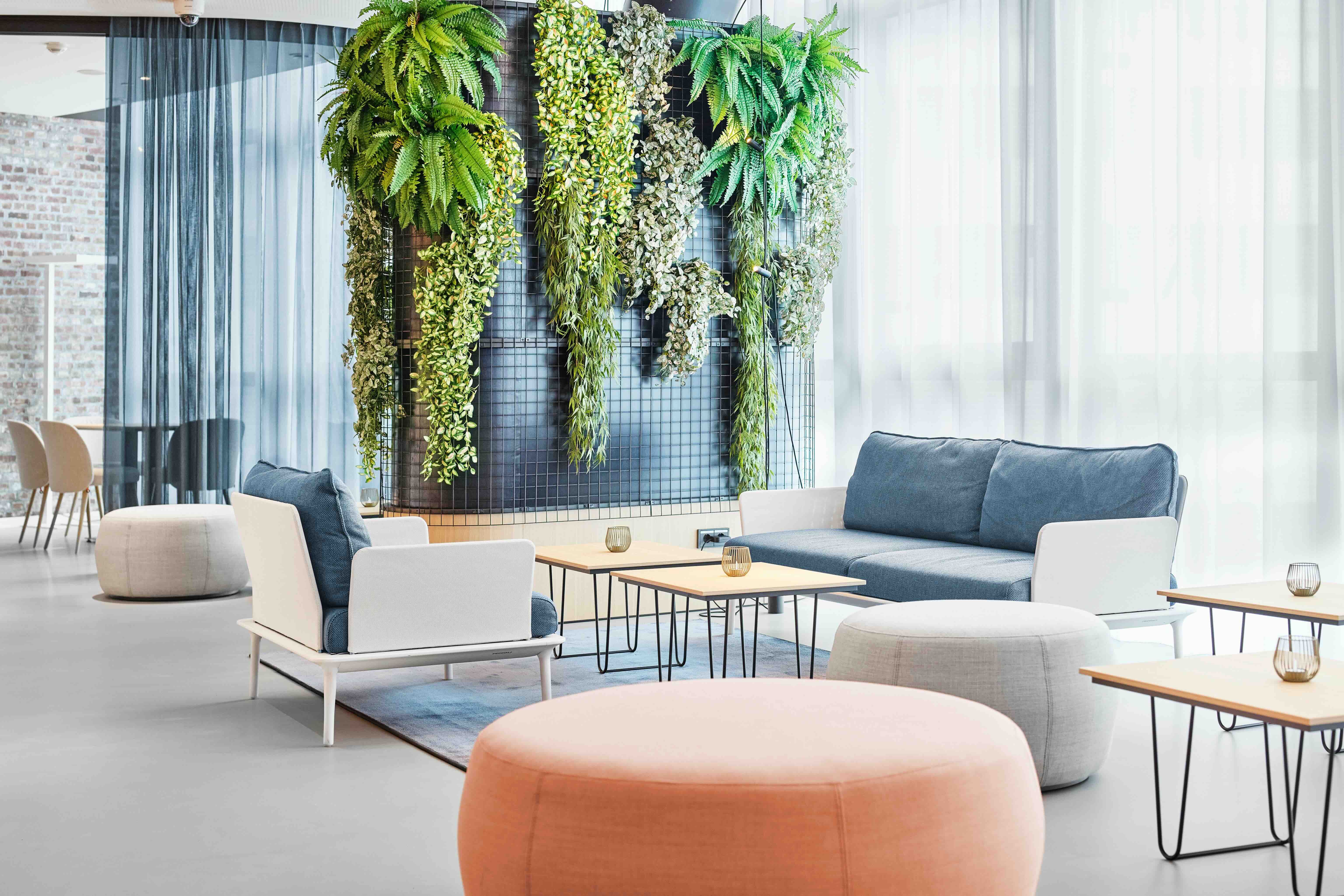 Modern eingerichteter Loungebereich mit bequemen Sitzmöbeln, hellen Farben und einer grünen Pflanzenwand, die eine entspannte und einladende Atmosphäre schafft. Große Fenster lassen viel Tageslicht herein.