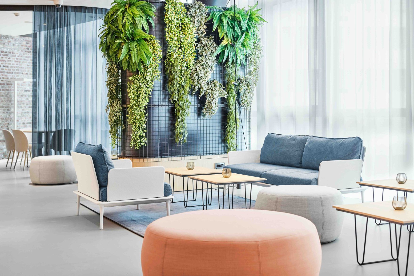 Modern eingerichteter Loungebereich mit bequemen Sitzmöbeln, hellen Farben und einer grünen Pflanzenwand, die eine entspannte und einladende Atmosphäre schafft. Große Fenster lassen viel Tageslicht herein.