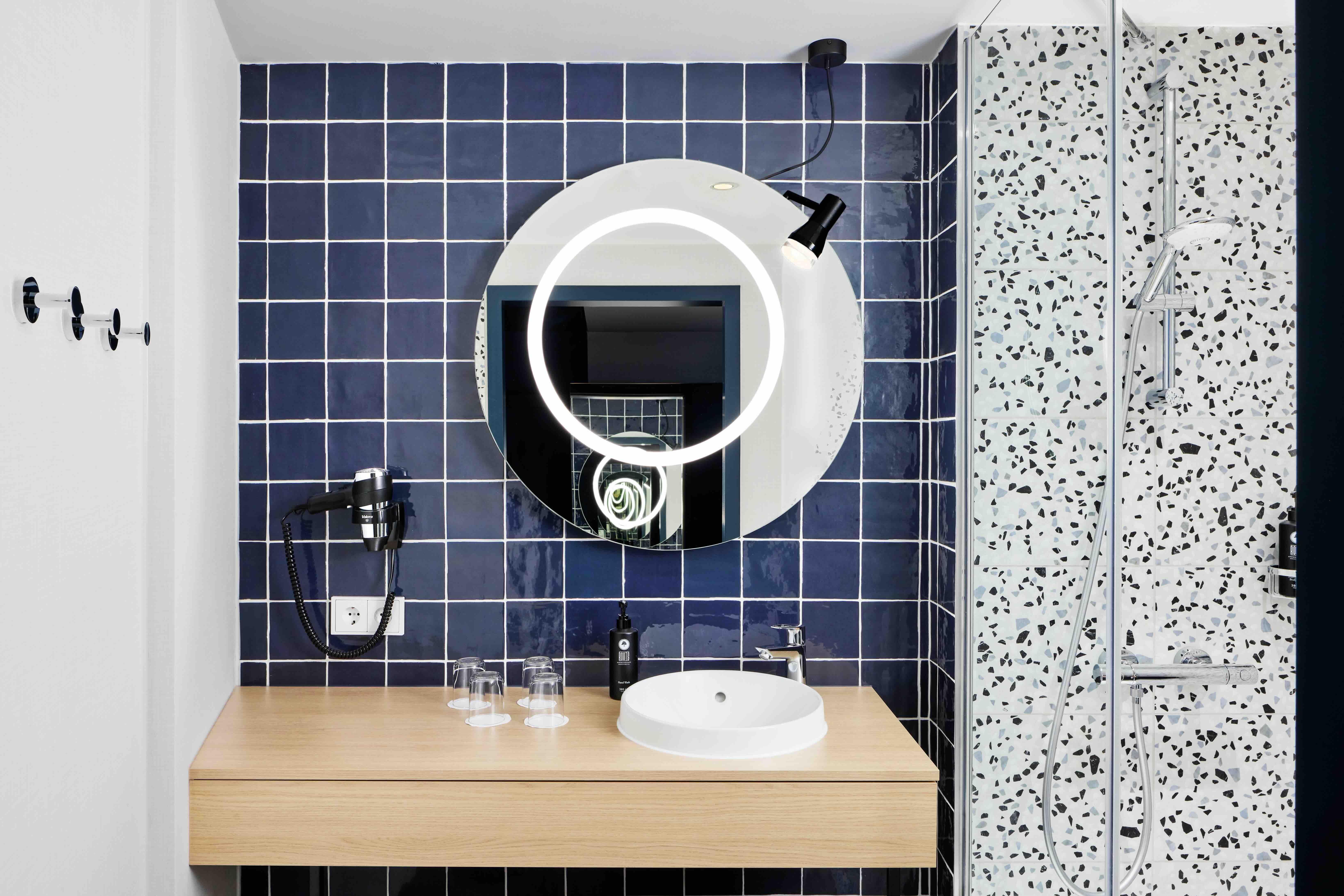 Modernes Badezimmer mit dunkelblauen Fliesen, rundem beleuchtetem Spiegel, weißem Waschbecken auf Holzplatte, integrierter Dusche mit Glaswand und Haartrockner an der Wand.