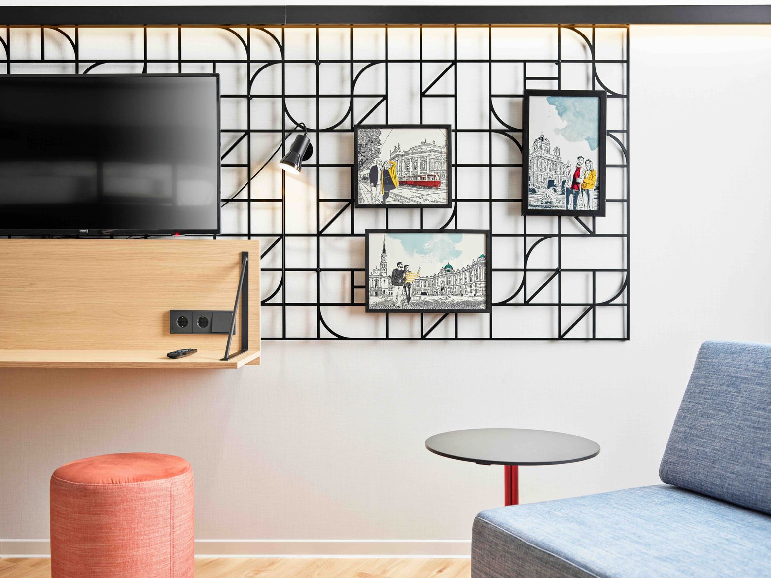 Moderner Hotelzimmerbereich mit flachem Fernseher, schwarzem Gitter-Wanddekor, drei städtischen Kunstdrucken, einem kleinen runden Tisch, einem blauen Sessel und einem orangefarbenen Hocker. Helles, stilvolles und funktionales Design.