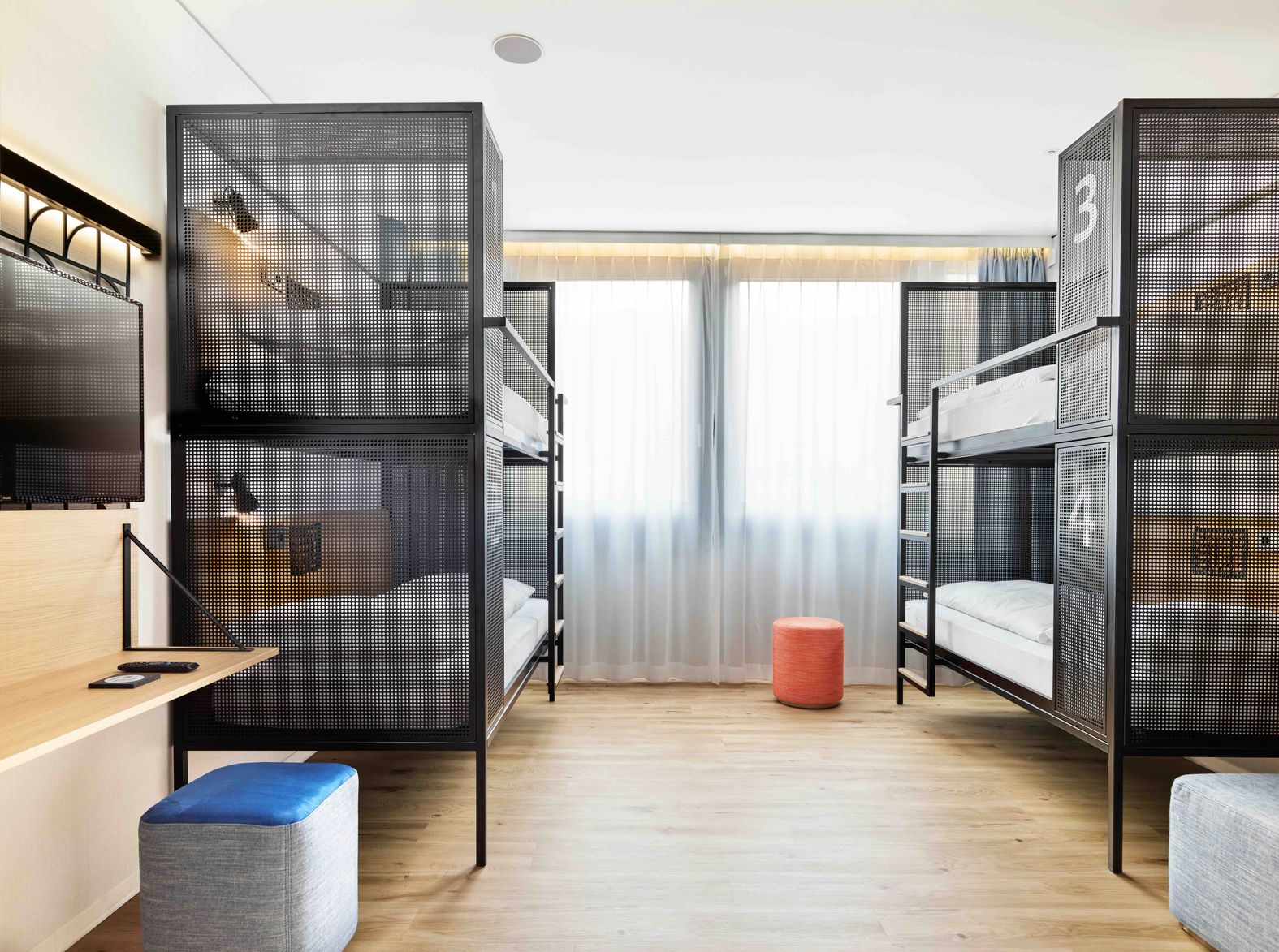 Moderne Vierbettzimmer mit zwei Etagenbetten, hellem Holzfußboden und großem Fenster für Tageslicht. Schlichte, funktionale Ausstattung mit Fernseher, kleinen Sitzhockern und angenehmer Atmosphäre für Gruppenreisende oder Familien.