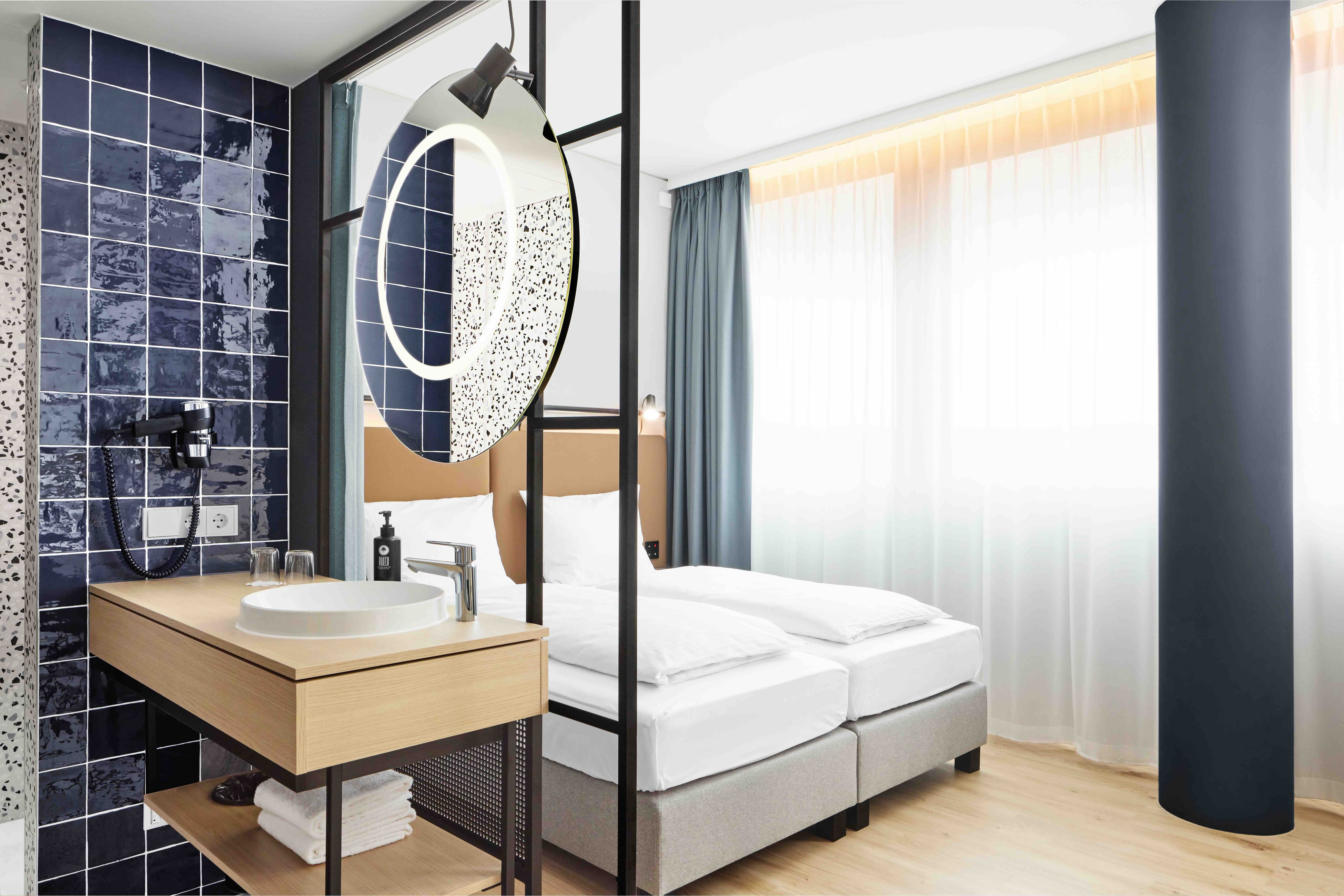 Modern gestaltetes Hotelzimmer mit gemütlichem Doppelbett, großzügigem Fenster mit Tageslicht und offenem Badezimmerbereich mit rundem Spiegel und stilvollem Waschbecken auf Hellholzmöbel.