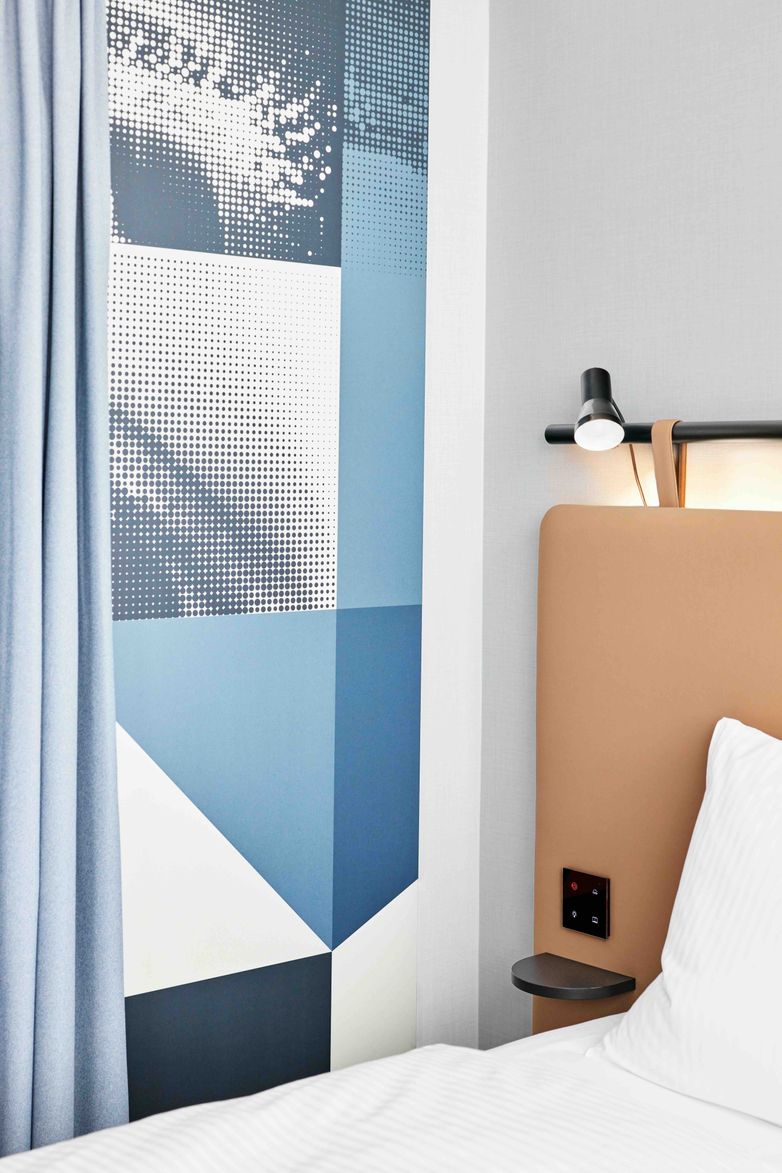Detailaufnahme eines modernen Hotelzimmers mit hellem Bett, gepolstertem Kopfteil, Leselampe und abstrakter Wandgestaltung in Blau- und Grautönen.