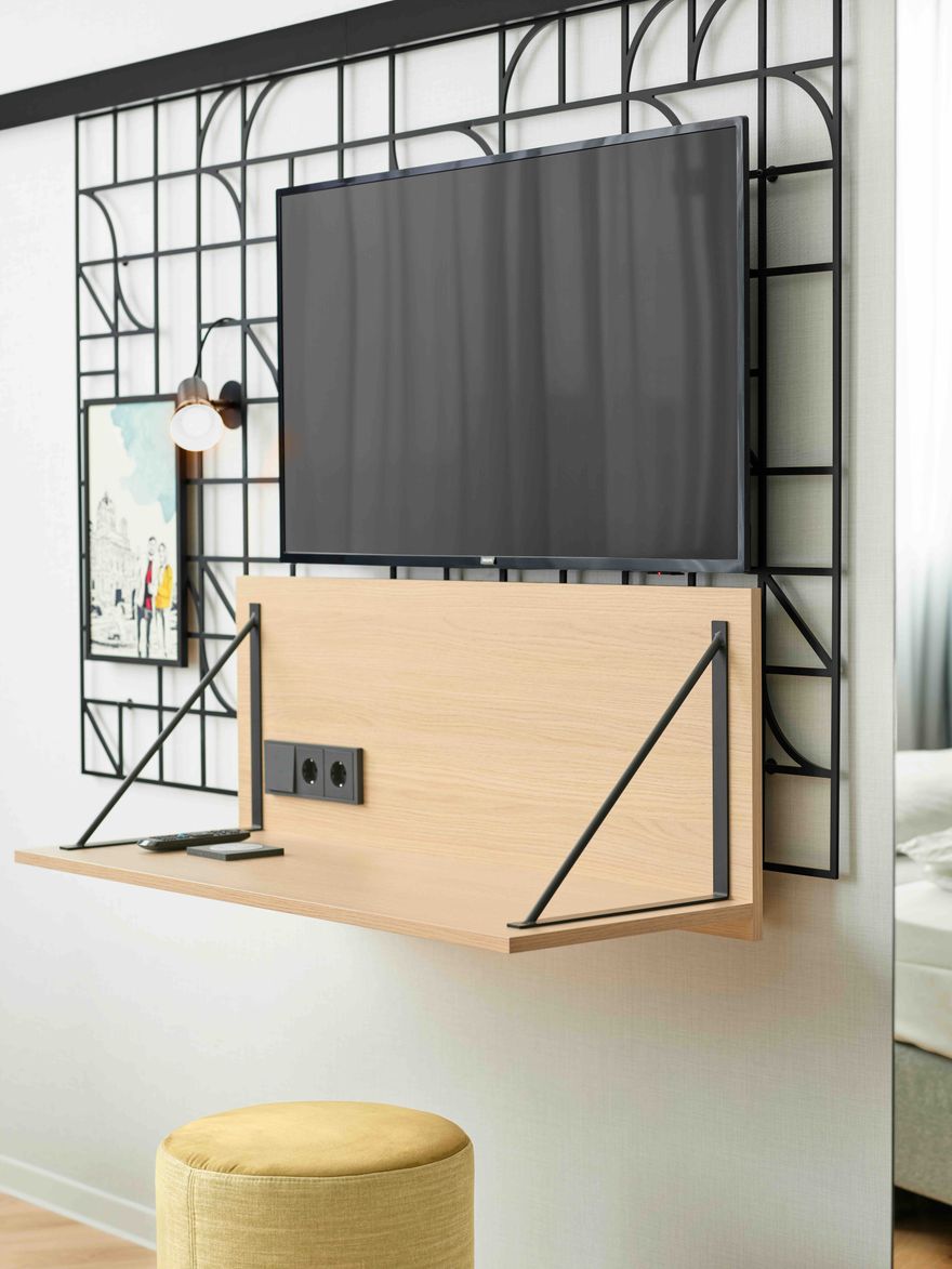 Moderner Arbeitsplatz mit klappbarem Holzregal unter einem Wandfernseher, integrierter Steckdose und stilvollem Gitterrahmen. Dazu ein bequemer, gelber Hocker – ideal für Arbeit oder Entspannung im Zimmer.