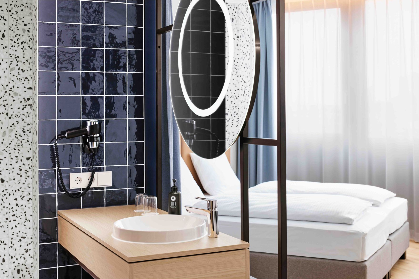 Modernes Hotelzimmer mit gemütlichem Bett, hellem Vorhang und Waschbecken auf Holzablage vor blauem Fliesenspiegel, ergänzt durch großen, runden Spiegel mit Beleuchtung für angenehme Atmosphäre.