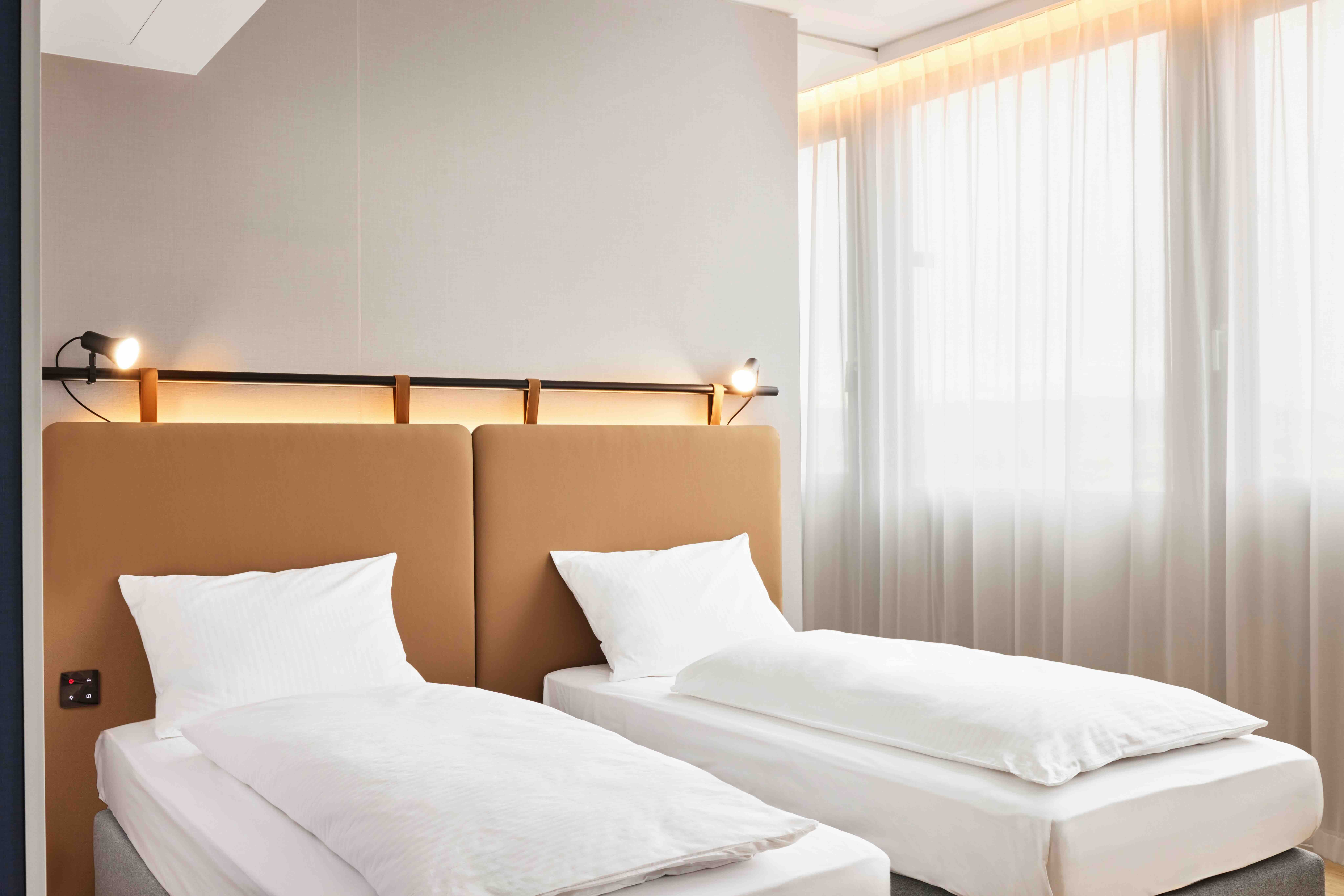 Zwei Einzelbetten mit weißer Bettwäsche in einem hellen, modernen Zimmer, großes Fenster mit lichtdurchlässigen Vorhängen, warme Beleuchtung und schlichte, einladende Atmosphäre.