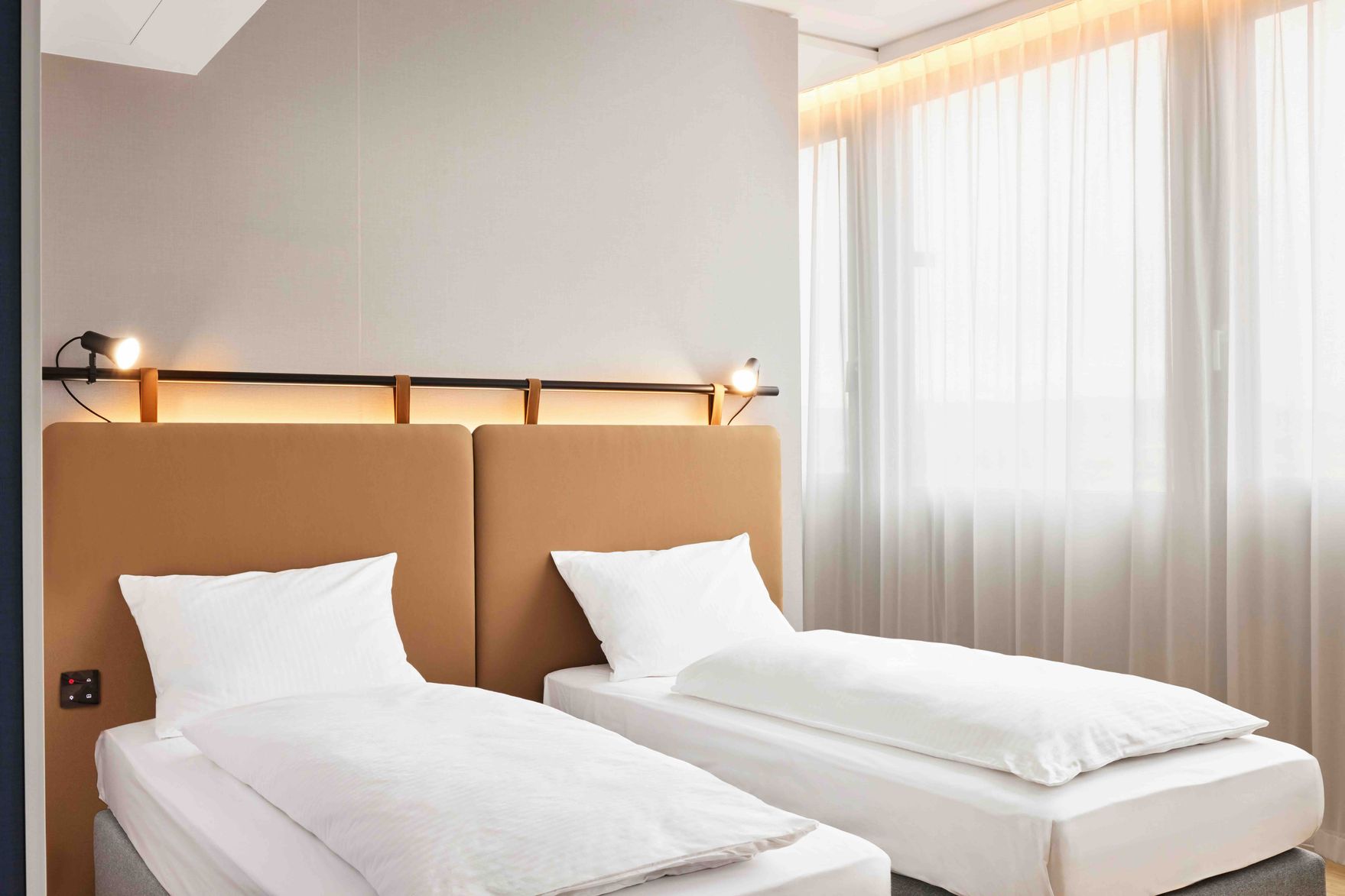Zwei Einzelbetten mit weißer Bettwäsche in einem hellen, modernen Zimmer, großes Fenster mit lichtdurchlässigen Vorhängen, warme Beleuchtung und schlichte, einladende Atmosphäre.