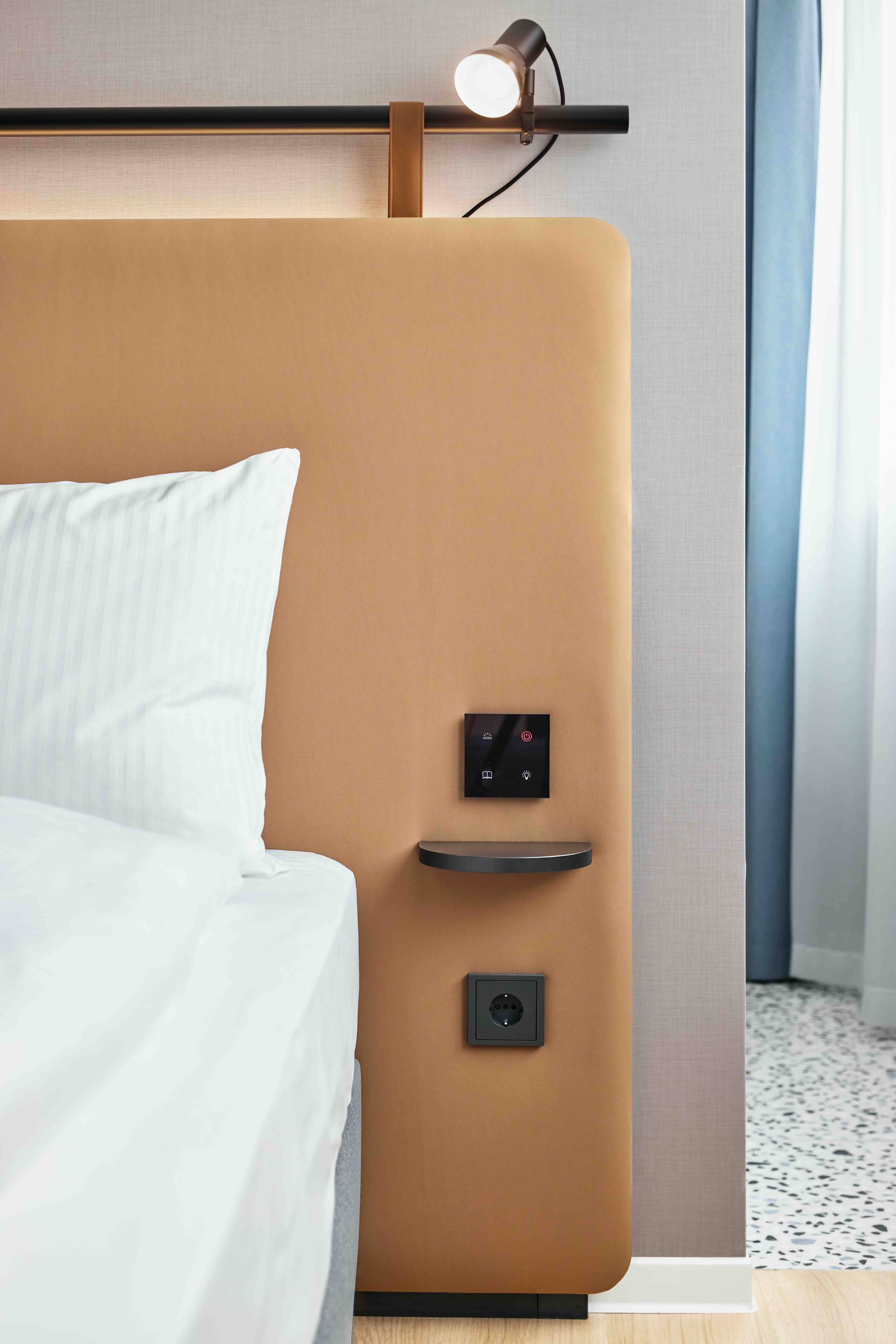 Detailaufnahme eines modernen Hotelbett-Kopfteil mit integriertem Licht, Nachtlichtschalter und Steckdose, helle Bettwäsche und Vorhänge vermitteln eine ruhige, komfortable Atmosphäre.